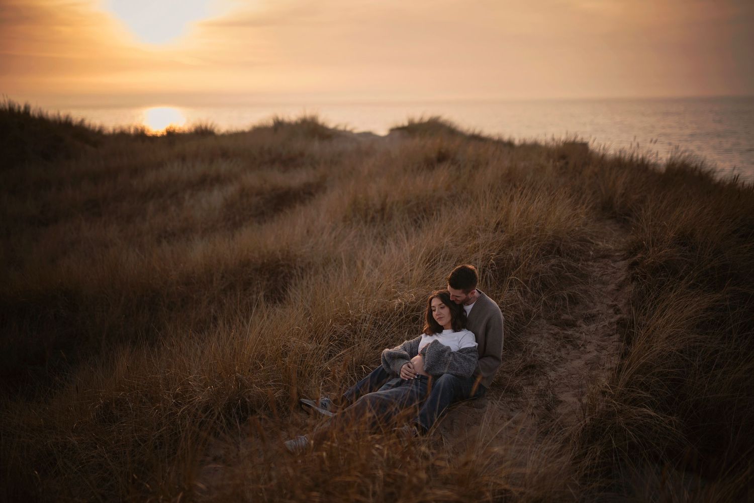 A couple sits together on a grassy coastal hillside at sunset while overlooking the ocean.