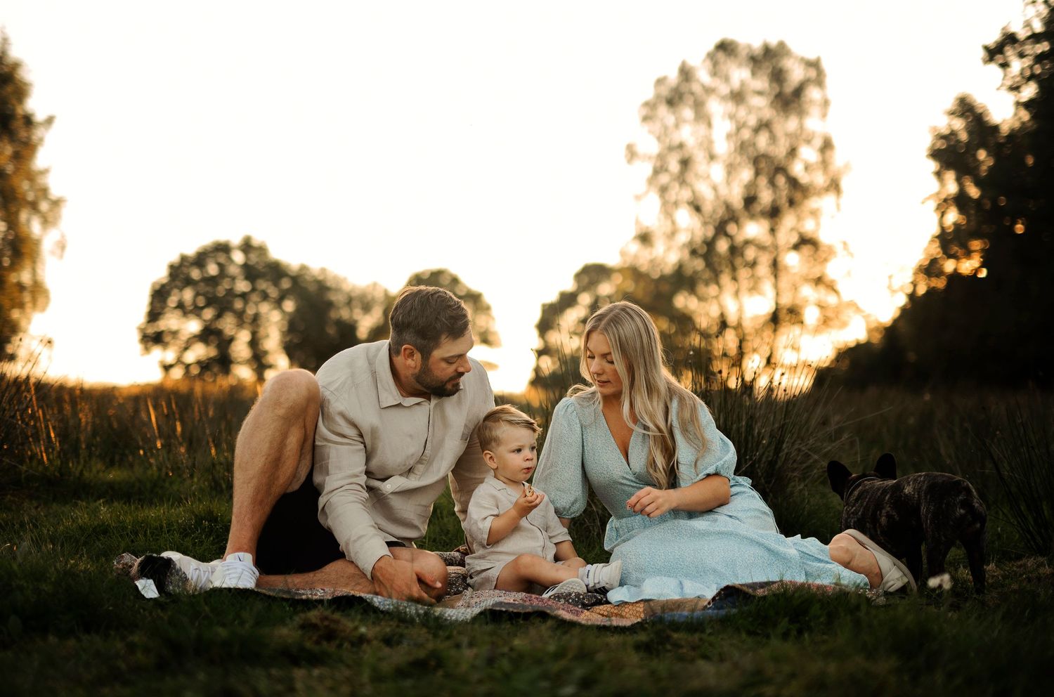 A family picnic scene at sunset with a small child and dog on a grassy hillside.
