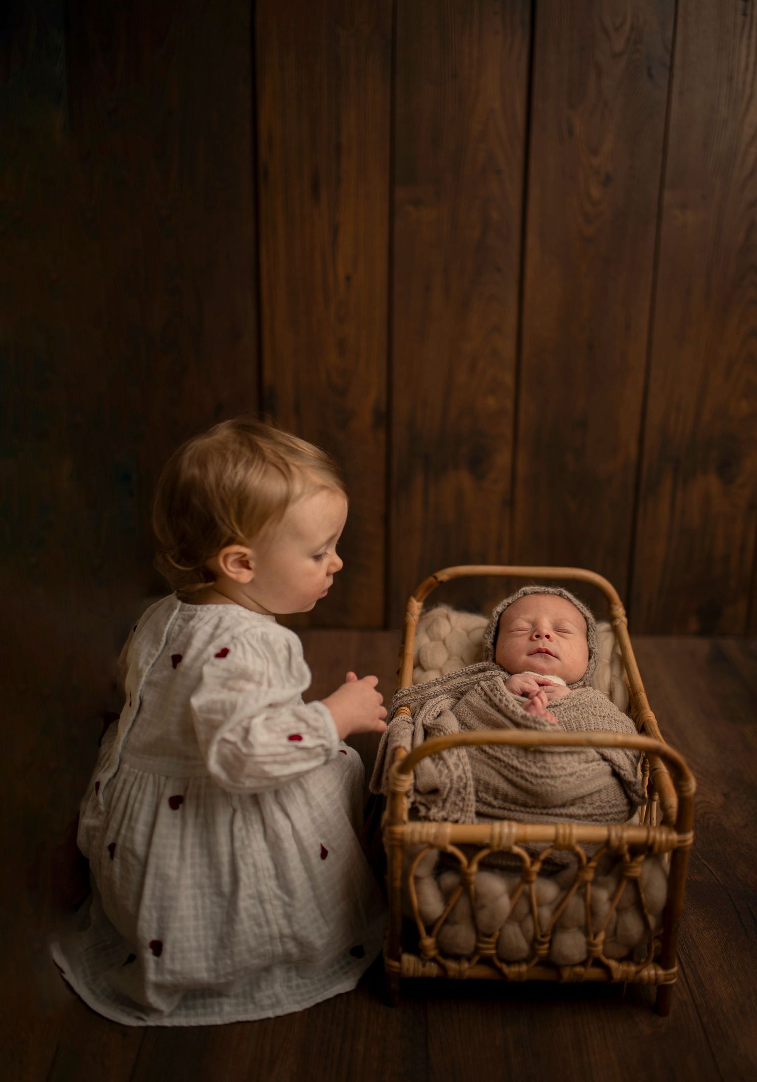 Toddler in white dress looking at baby sibling in vintage wicker bassinet against wooden wall.
