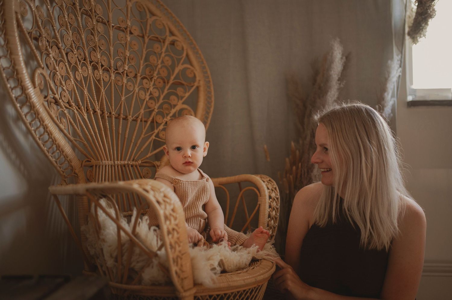 Joyful interaction between parent and baby seated by a peacock chair with natural lighting.