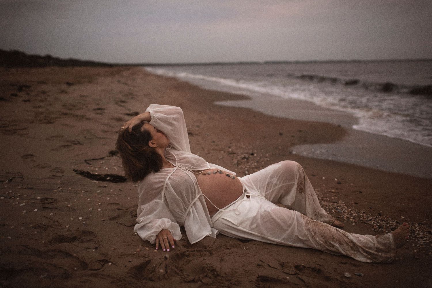 Figure in flowing white dress reclines on beach sand during dreamy sunset photoshoot.