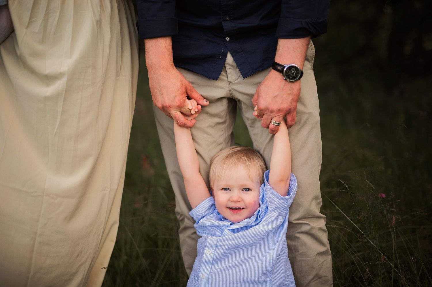 A small child in blue clothing holds onto adult hands while standing in a grassy outdoor setting.