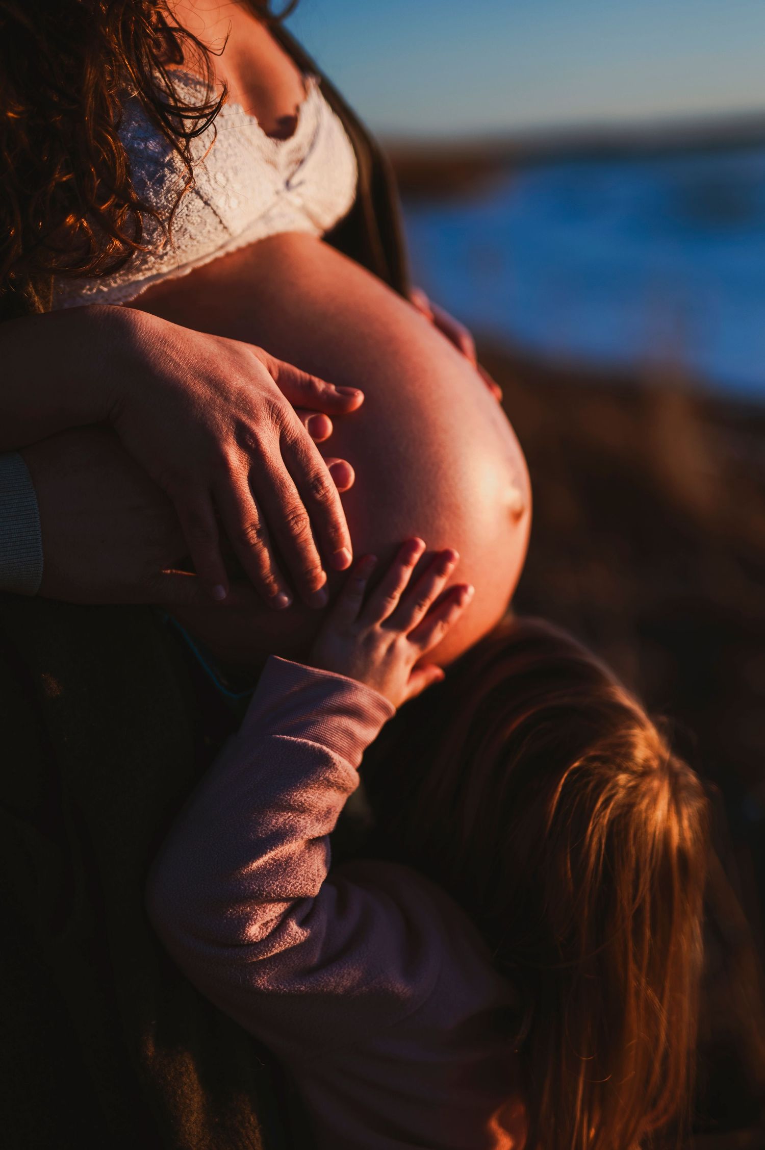 Small child's hands touching a pregnant belly at sunset by the ocean.