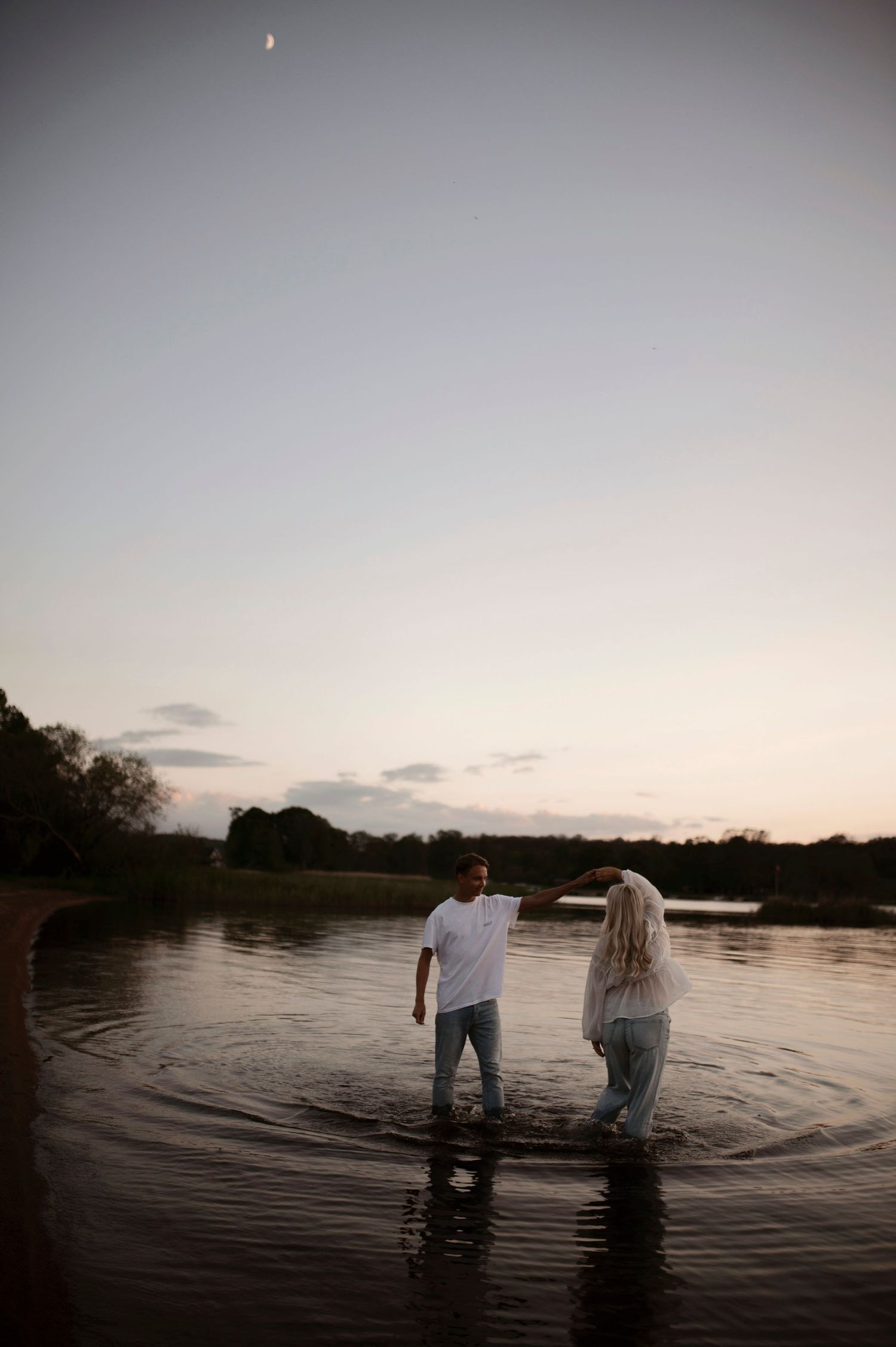 A couple wades in calm water at sunset with a crescent moon visible in the sky.