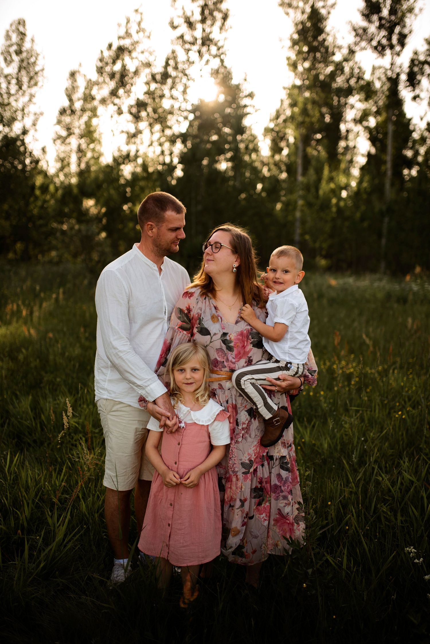 Family portrait in a sunlit field with tall trees in the background at golden hour.
