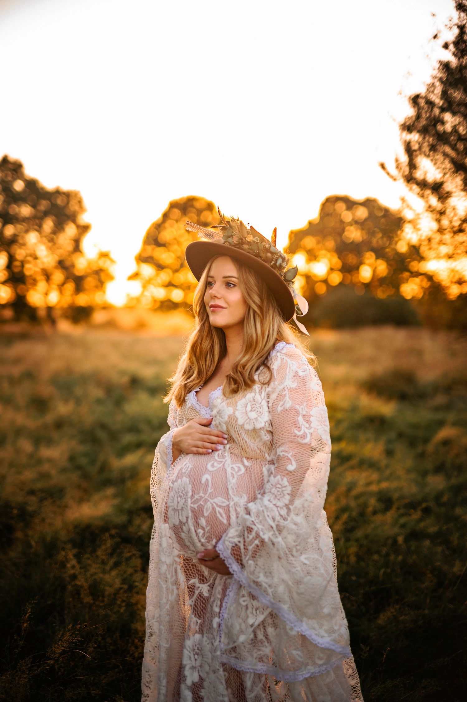Bohemian maternity photo featuring white lace dress and wide-brimmed hat at sunset.