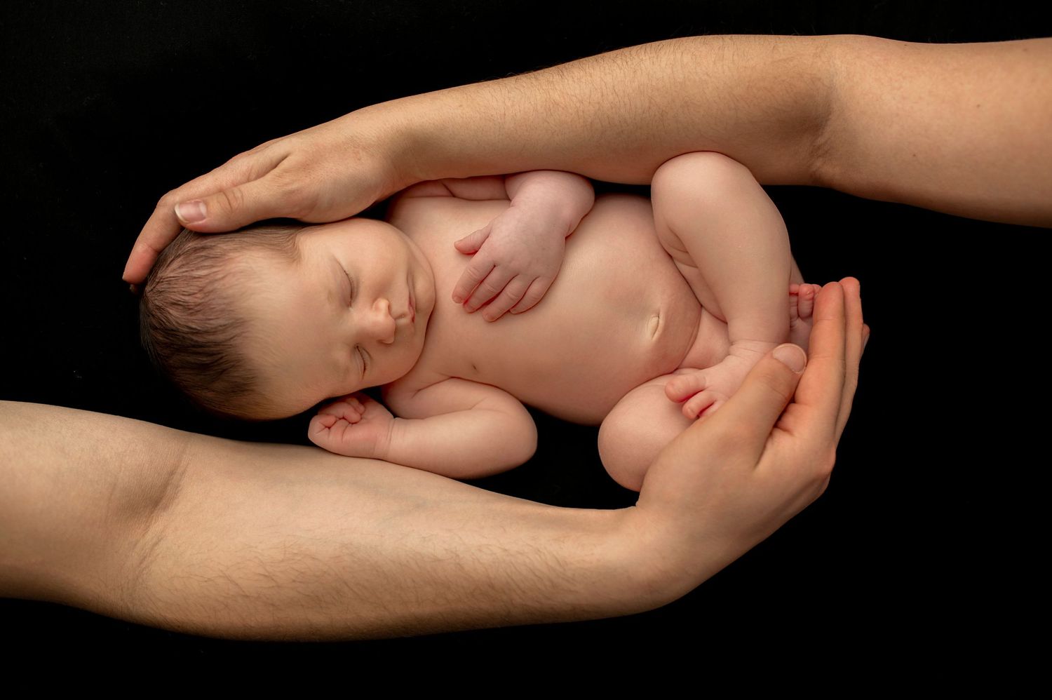 Sleeping newborn cradled in adult hands against black background in artistic portrait.