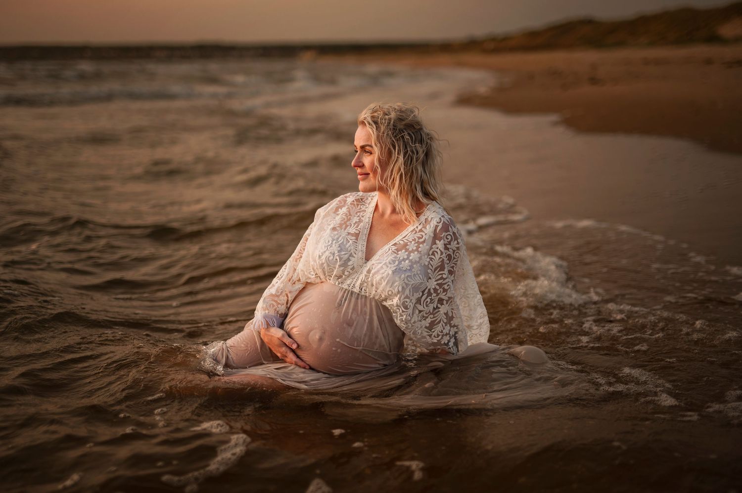 Pregnant woman in white lace dress sitting in ocean waves at sunset during a maternity photo session.