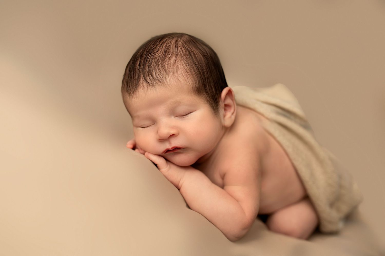 Peaceful newborn baby sleeping on beige backdrop with hands tucked under chin.