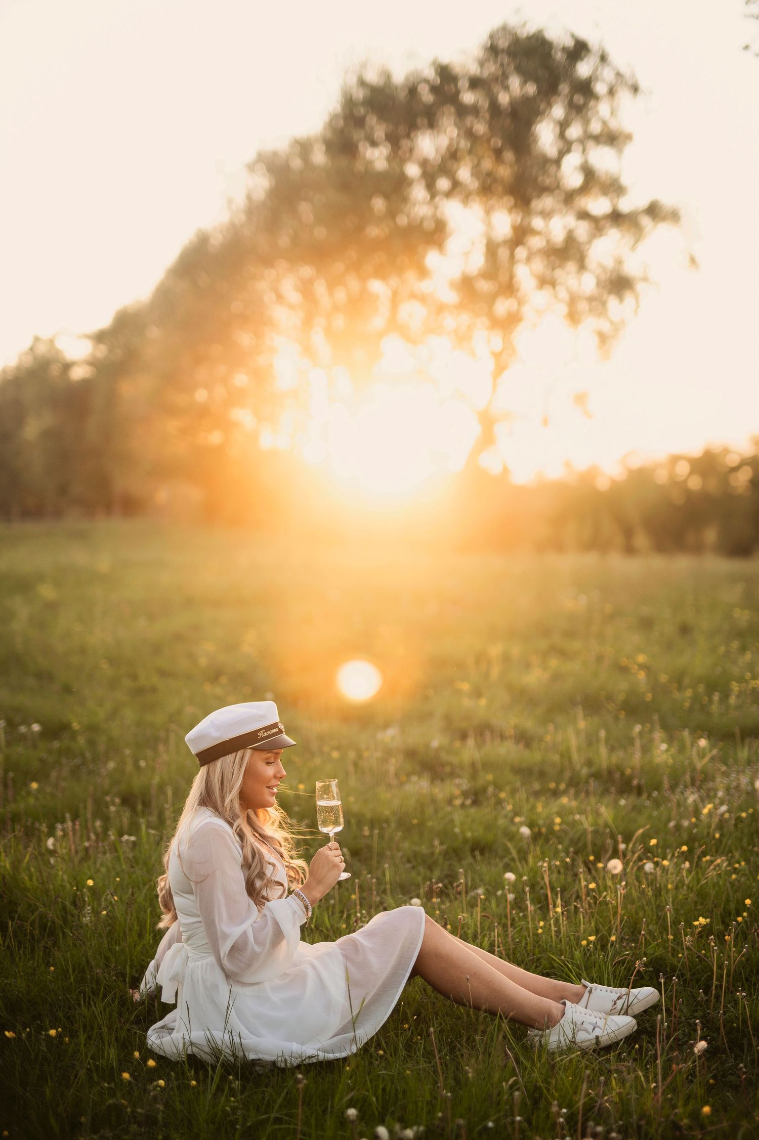 Blonde woman in white dress and captain hat holding champagne glass in sunny meadow.