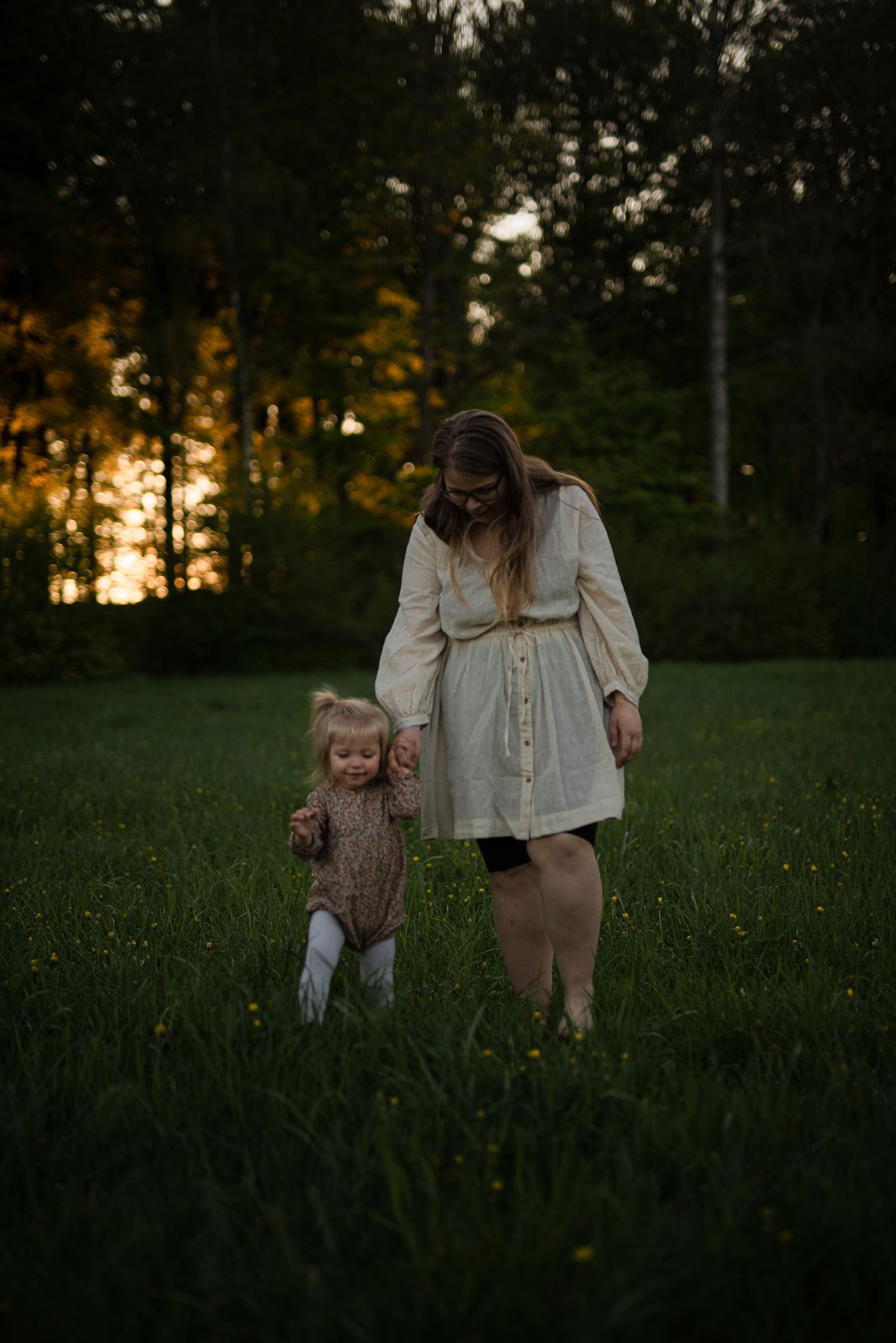A mother and toddler walk hand in hand through a green meadow at golden hour with trees in the background.