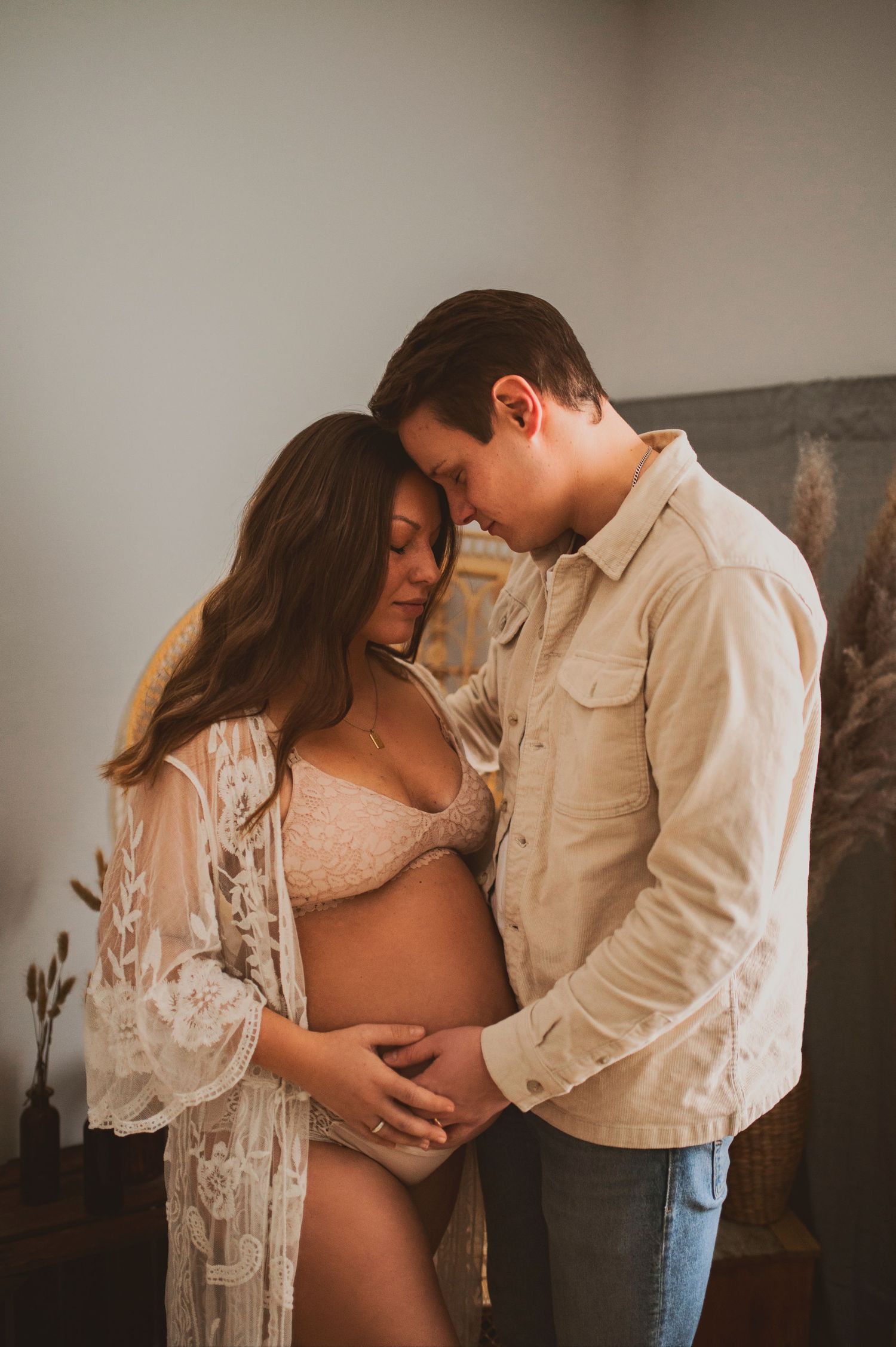 Intimate indoor maternity photo with couple sharing tender moment against neutral wall.
