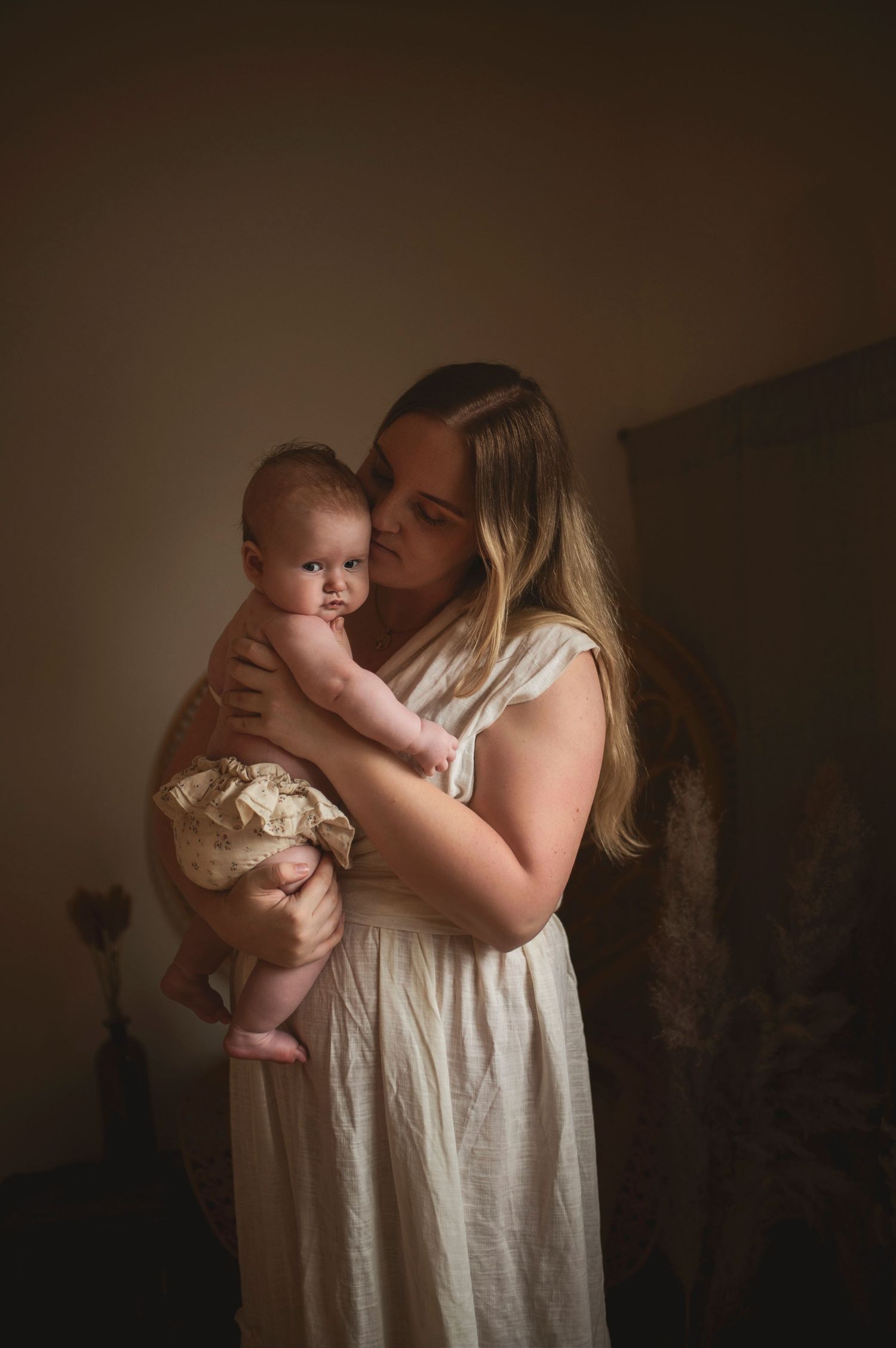Atmospheric portrait of parent holding baby in white dress against moody dark backdrop.