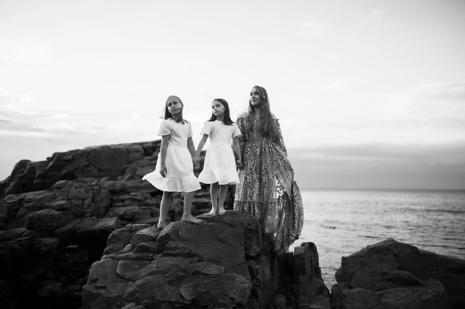 Three figures in light dresses stand together on rocky cliffs overlooking the ocean at sunset.