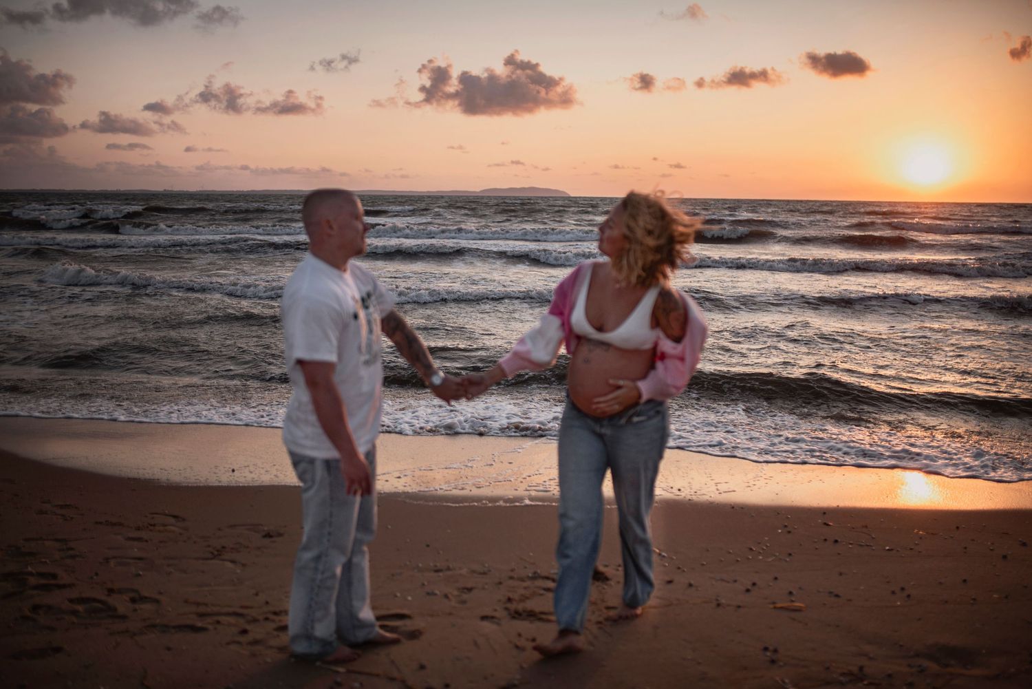 Two people dance together on a sandy beach at sunset with ocean waves crashing in the background.