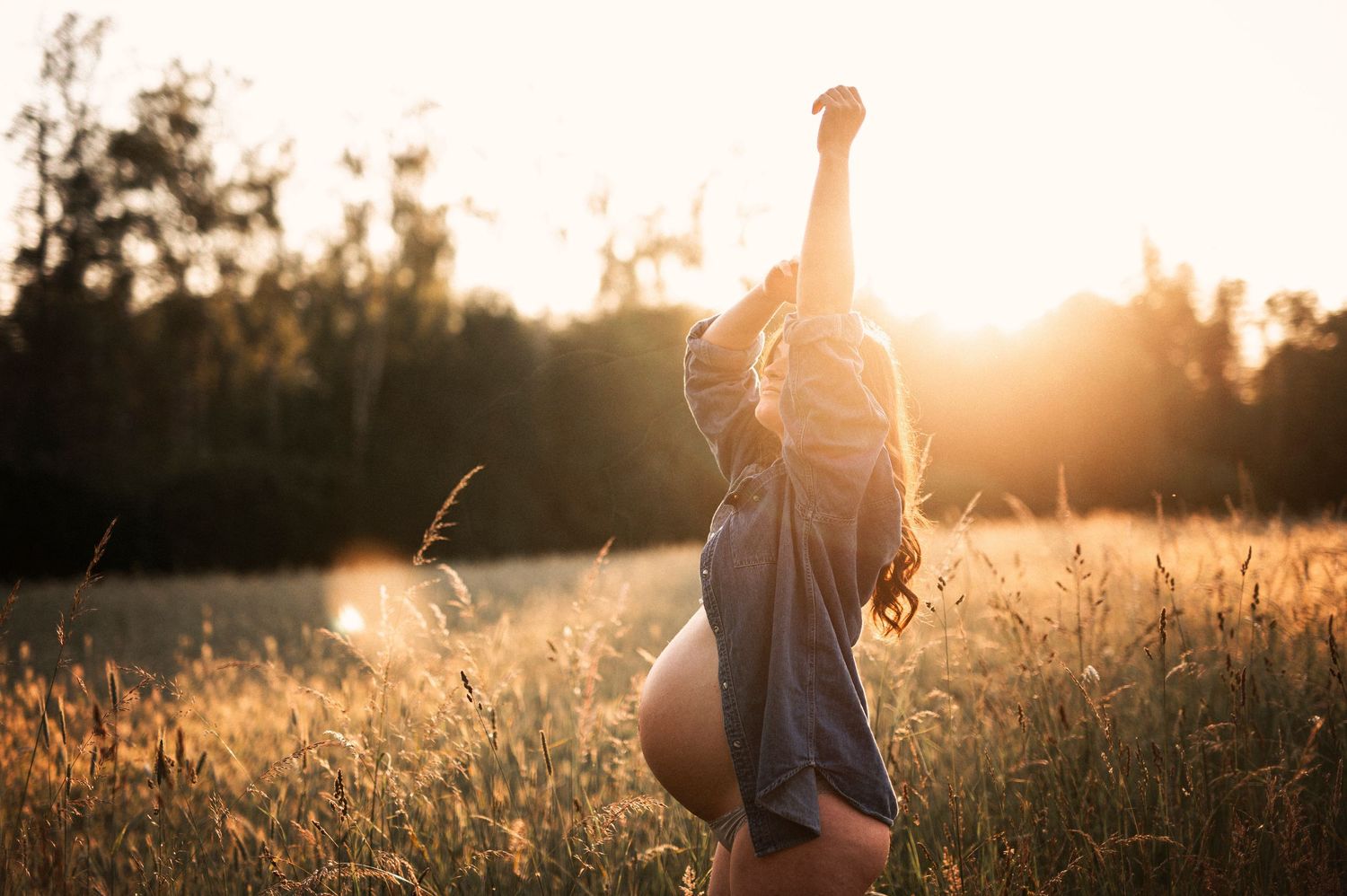 A silhouetted figure stands joyfully in a sunlit field with arms raised during golden hour.