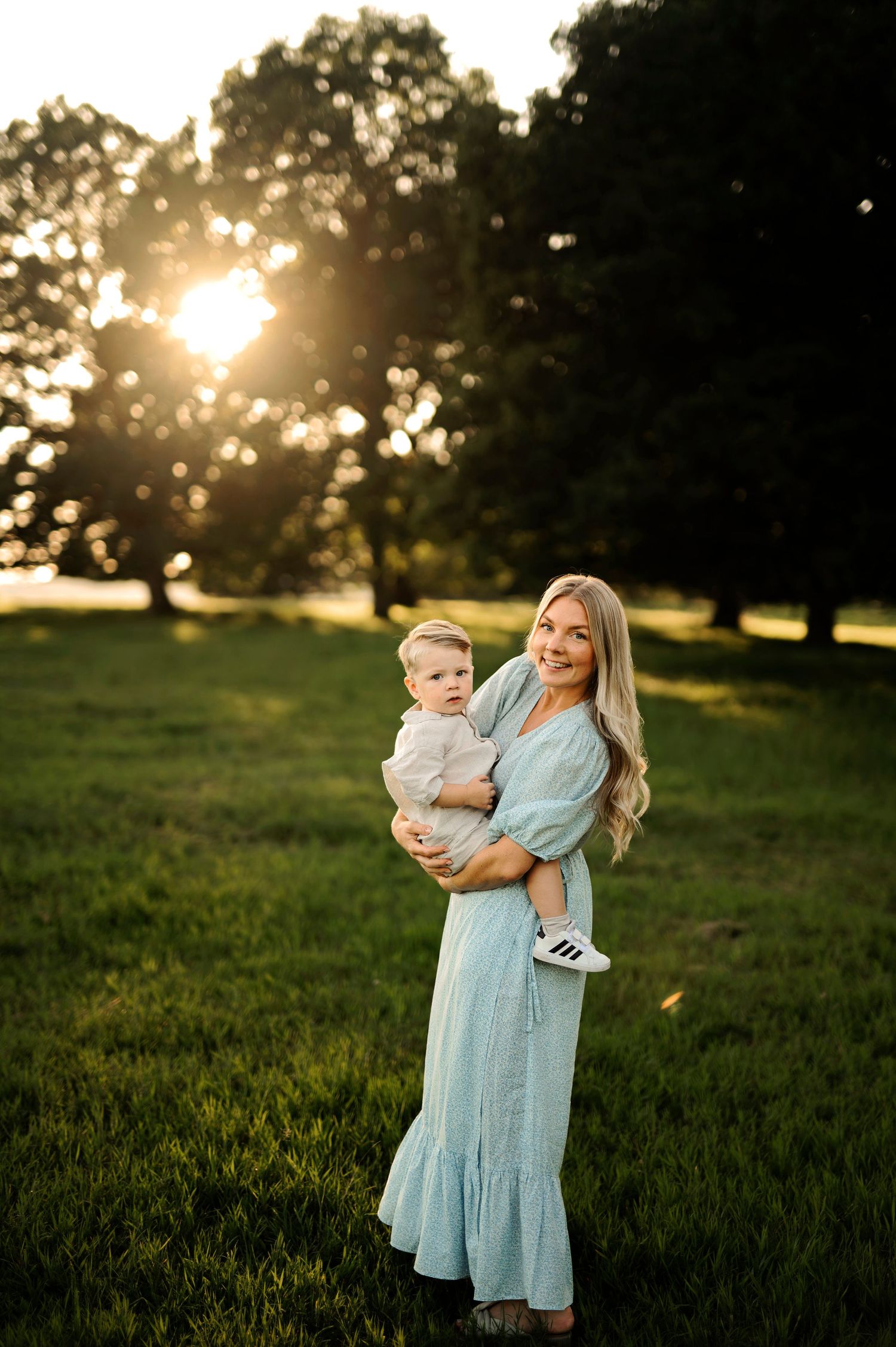 Mother in blue dress holds child while standing in sunlit grassy field at golden hour.