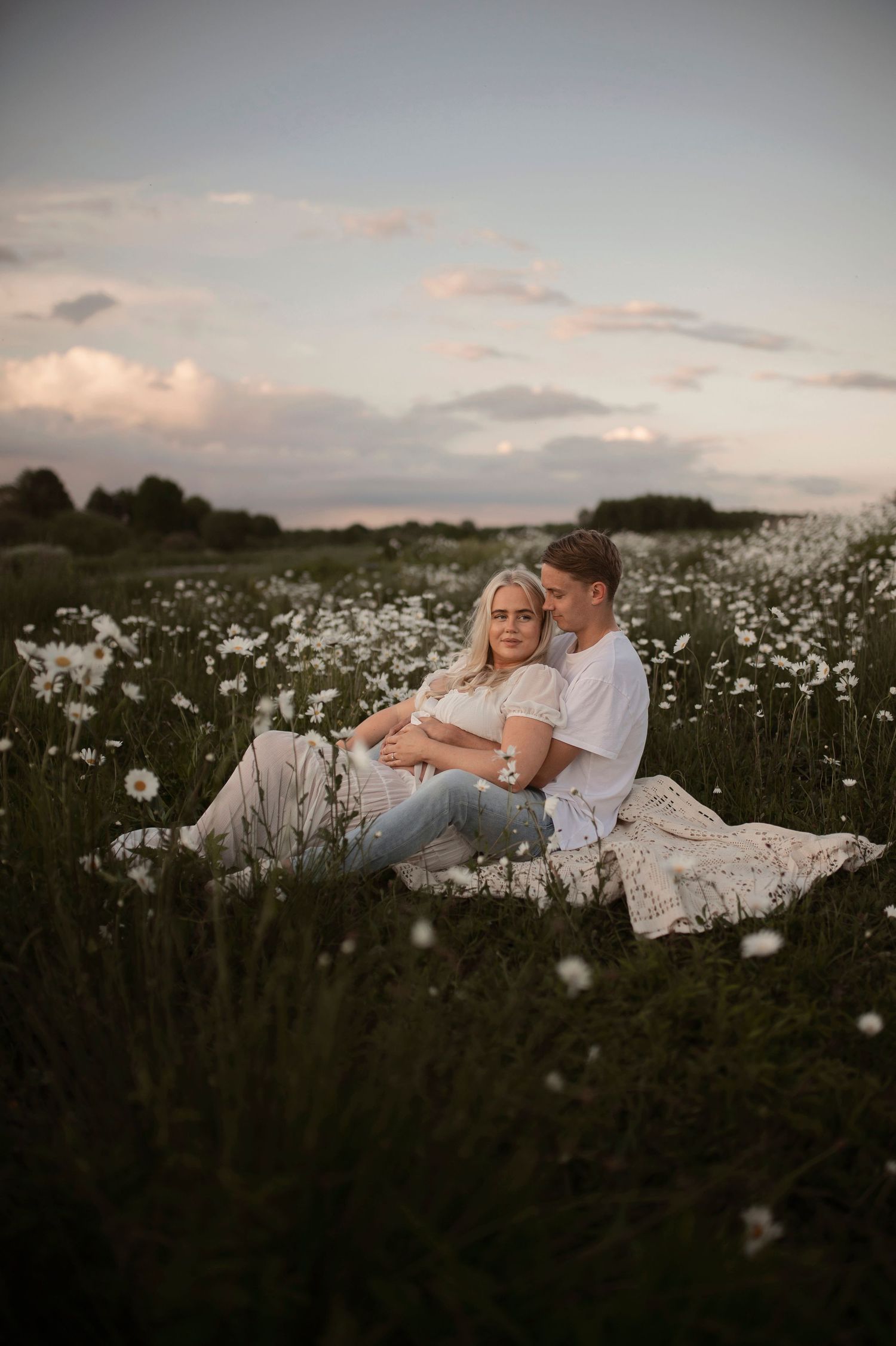 A couple relaxes together on a blanket in a dreamy meadow of white wildflowers at sunset.