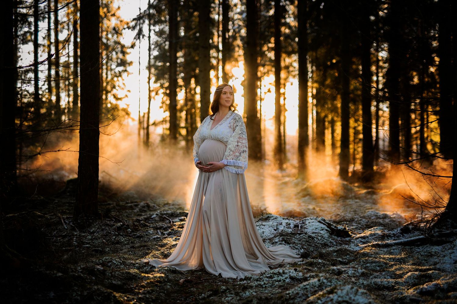 Pregnant woman in lace gown glowing in golden forest sunset light during maternity photoshoot.