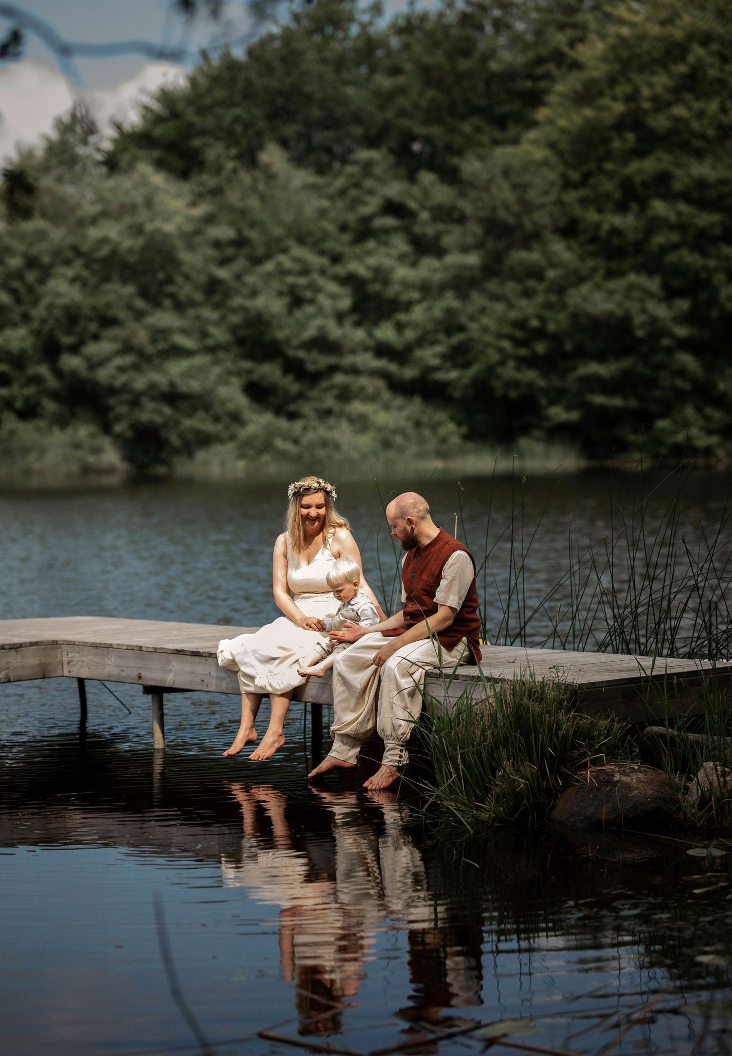Family with baby sitting on wooden dock by lake with feet in water.