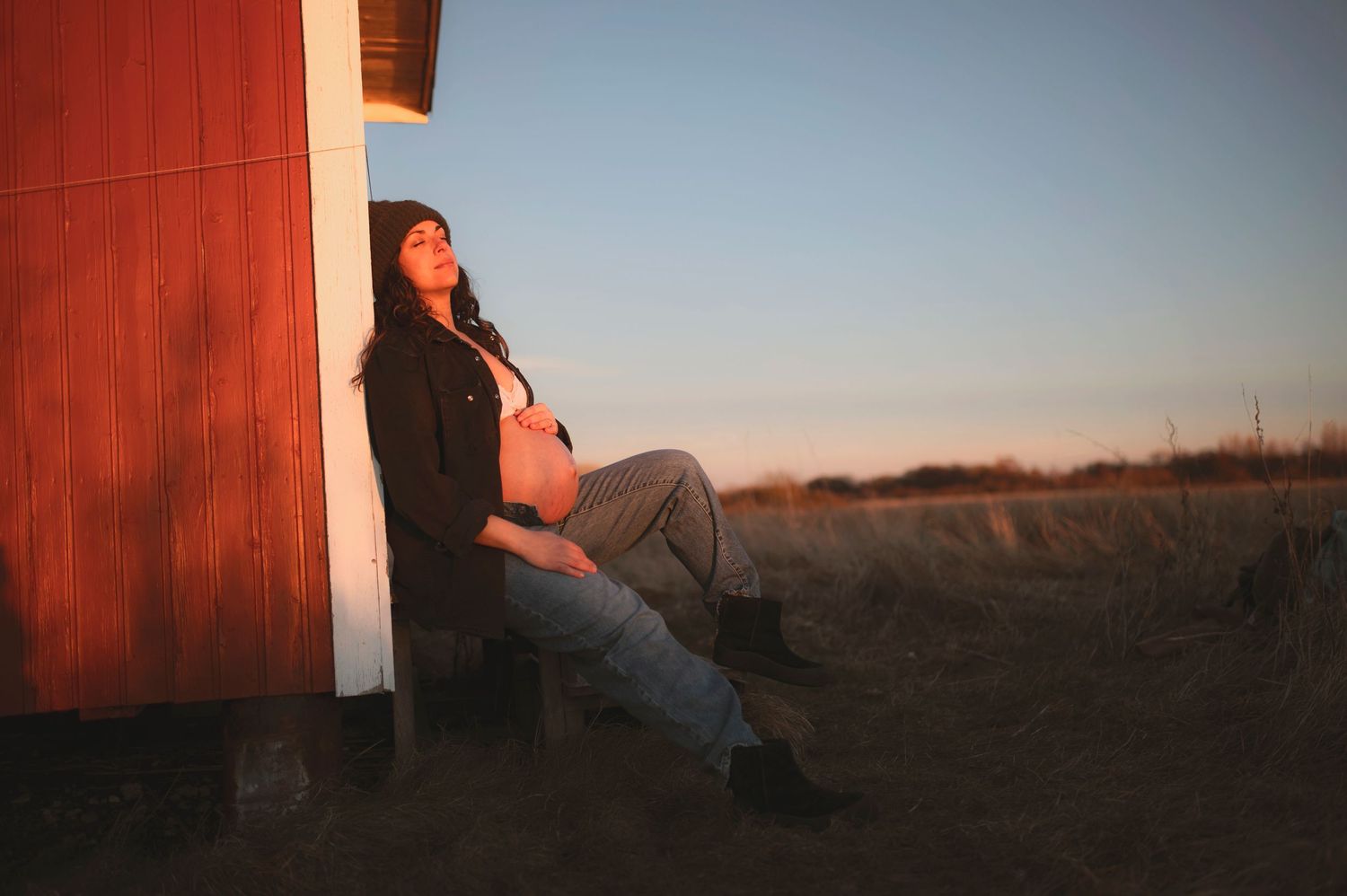 Maternity portrait at sunset against red wooden barn in rural setting.