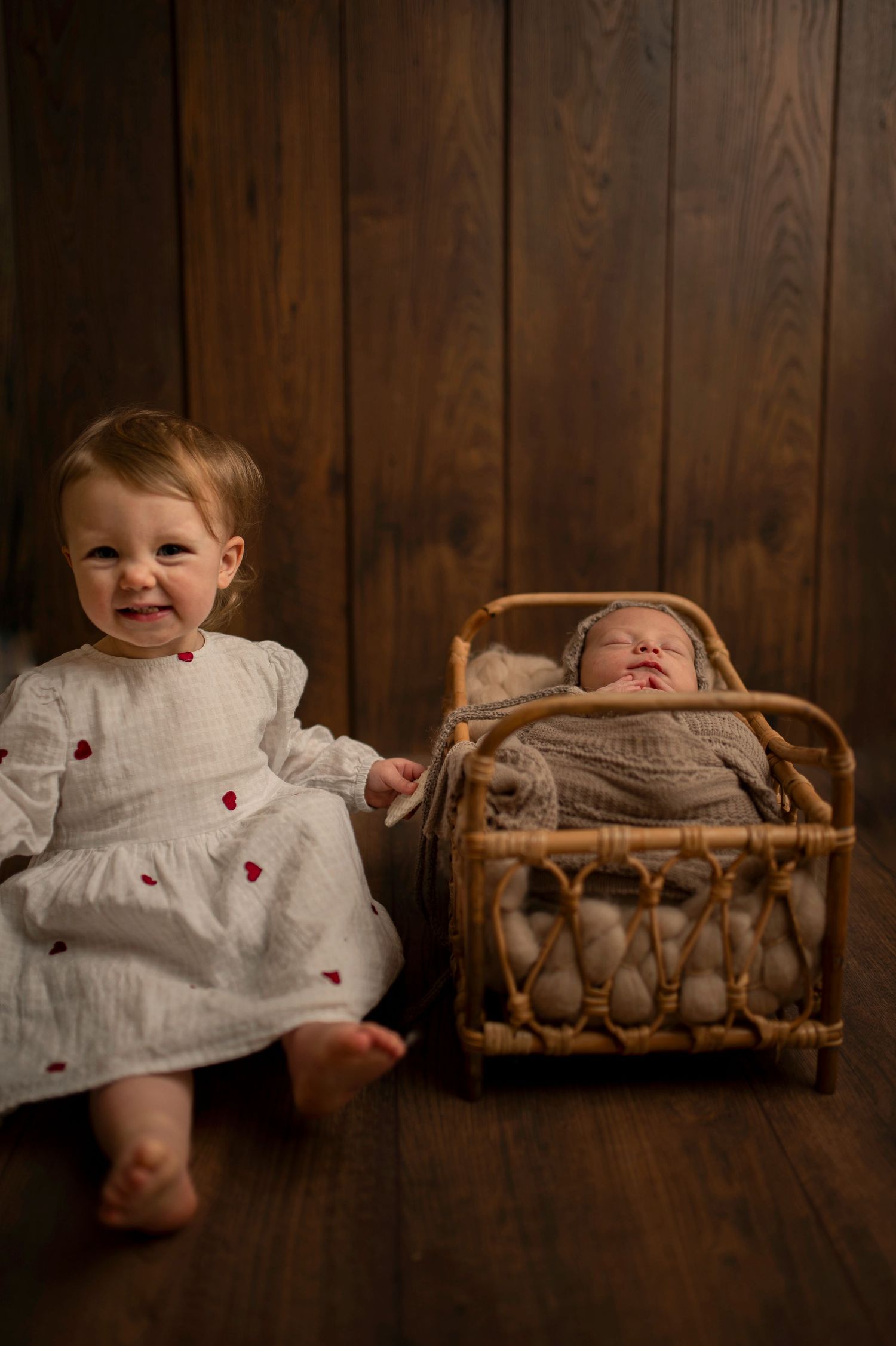 Two small children sit against a wooden wall, with one in a white dress and another in a wicker bassinet.
