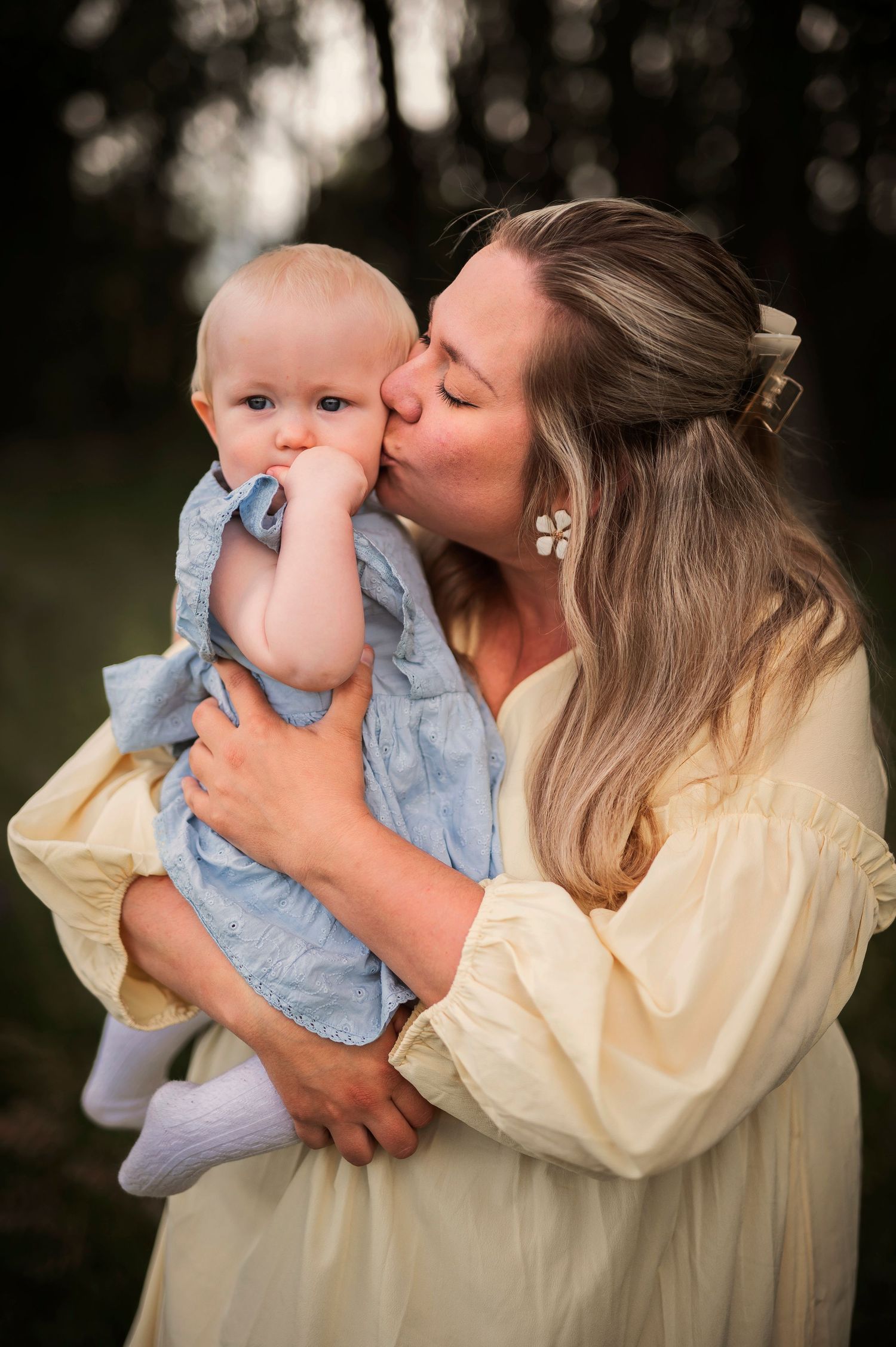 Tender moment between parent and baby in cream and blue outfits against a dark forest background.