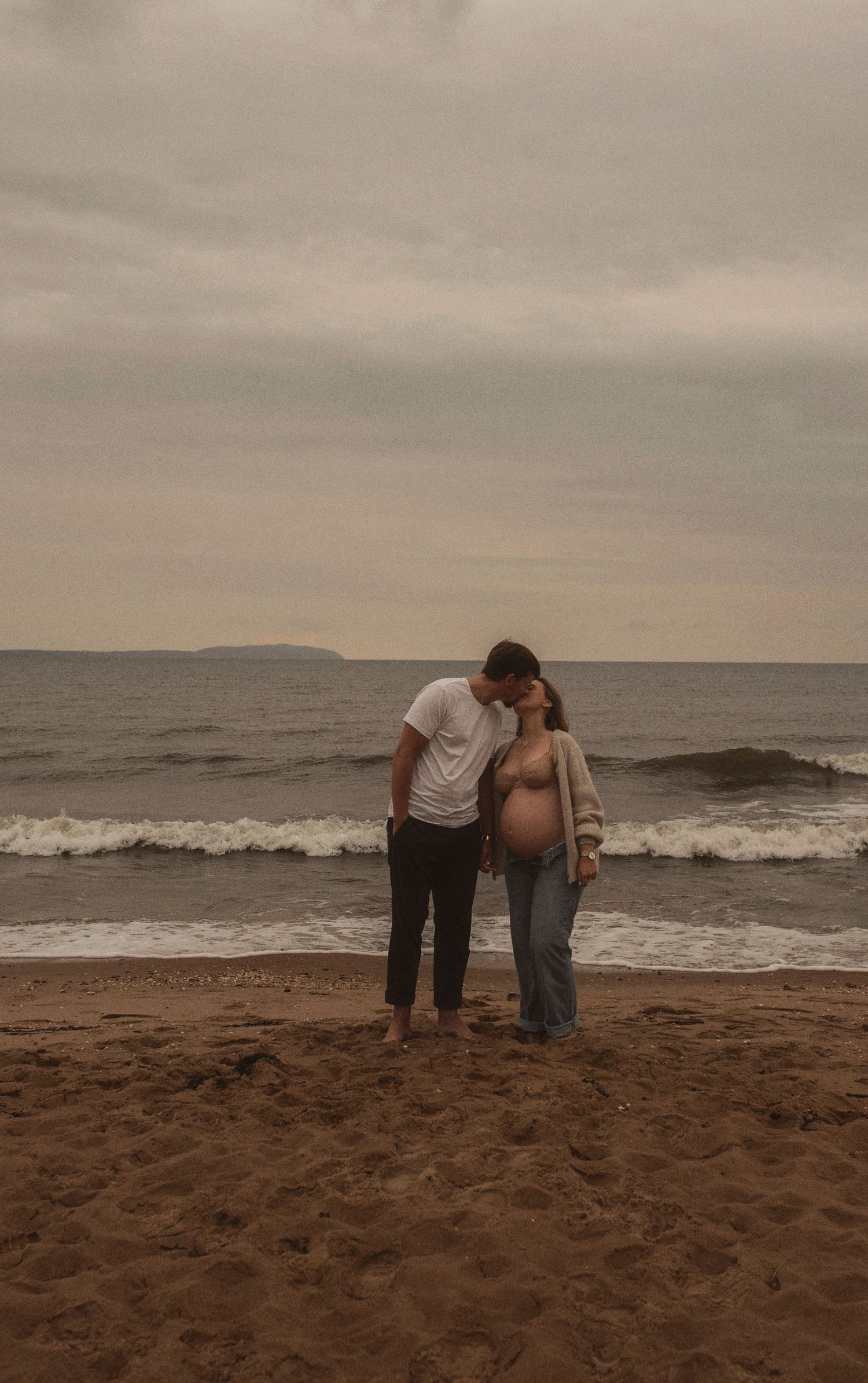 Couple stands together on sandy beach with overcast sky and rolling waves.