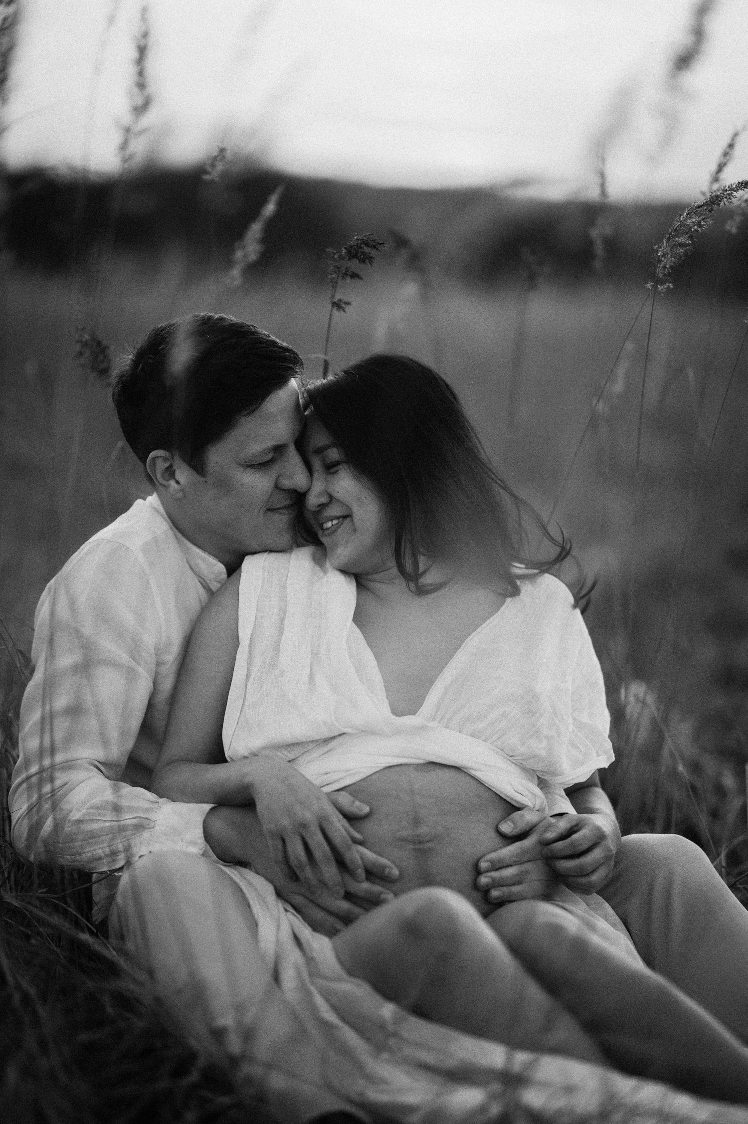 A romantic black and white maternity photo showing a couple in white clothing embracing while sitting in tall grass.