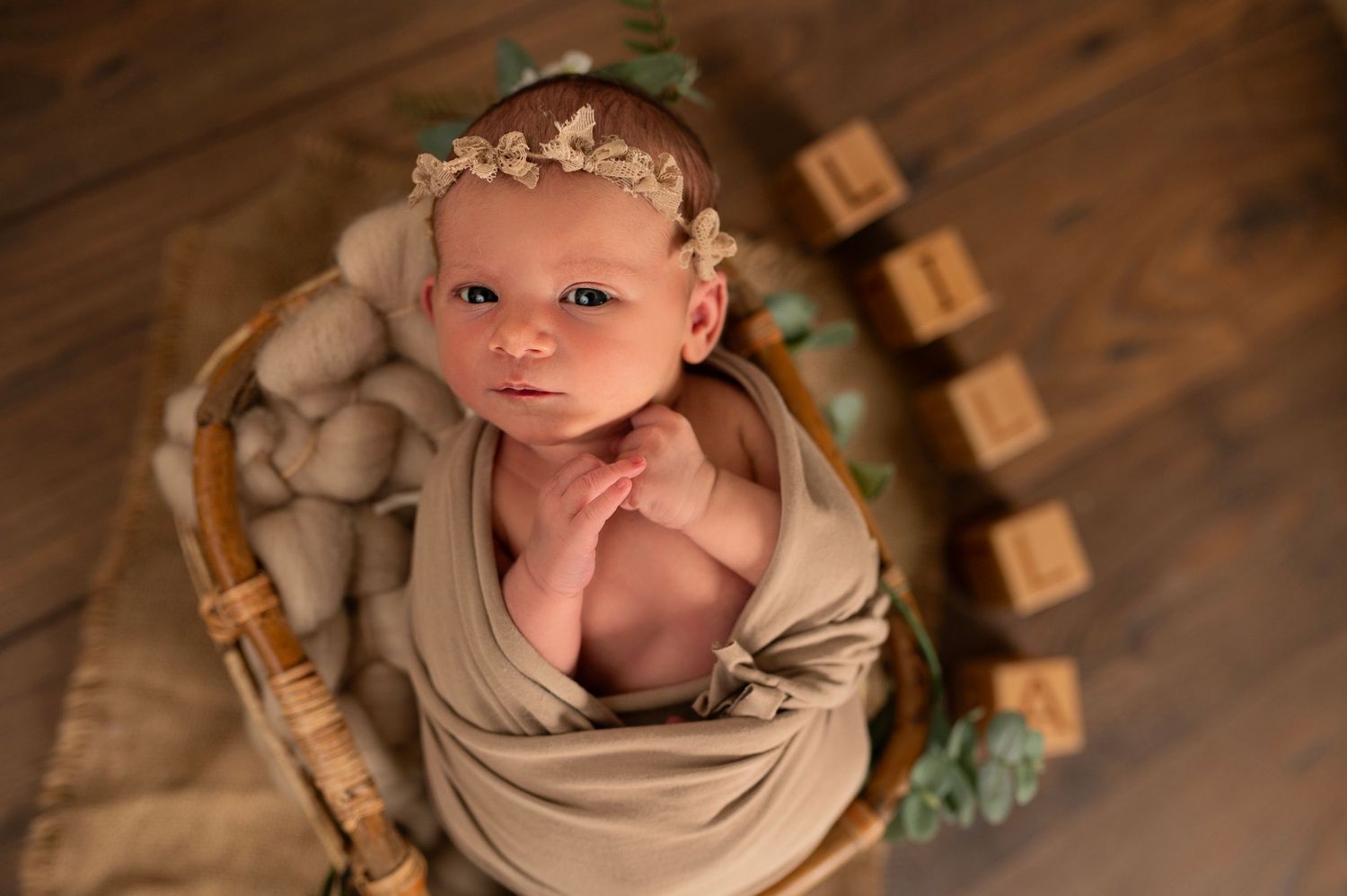 Newborn photography featuring baby wrapped in beige fabric with floral crown and wooden blocks.