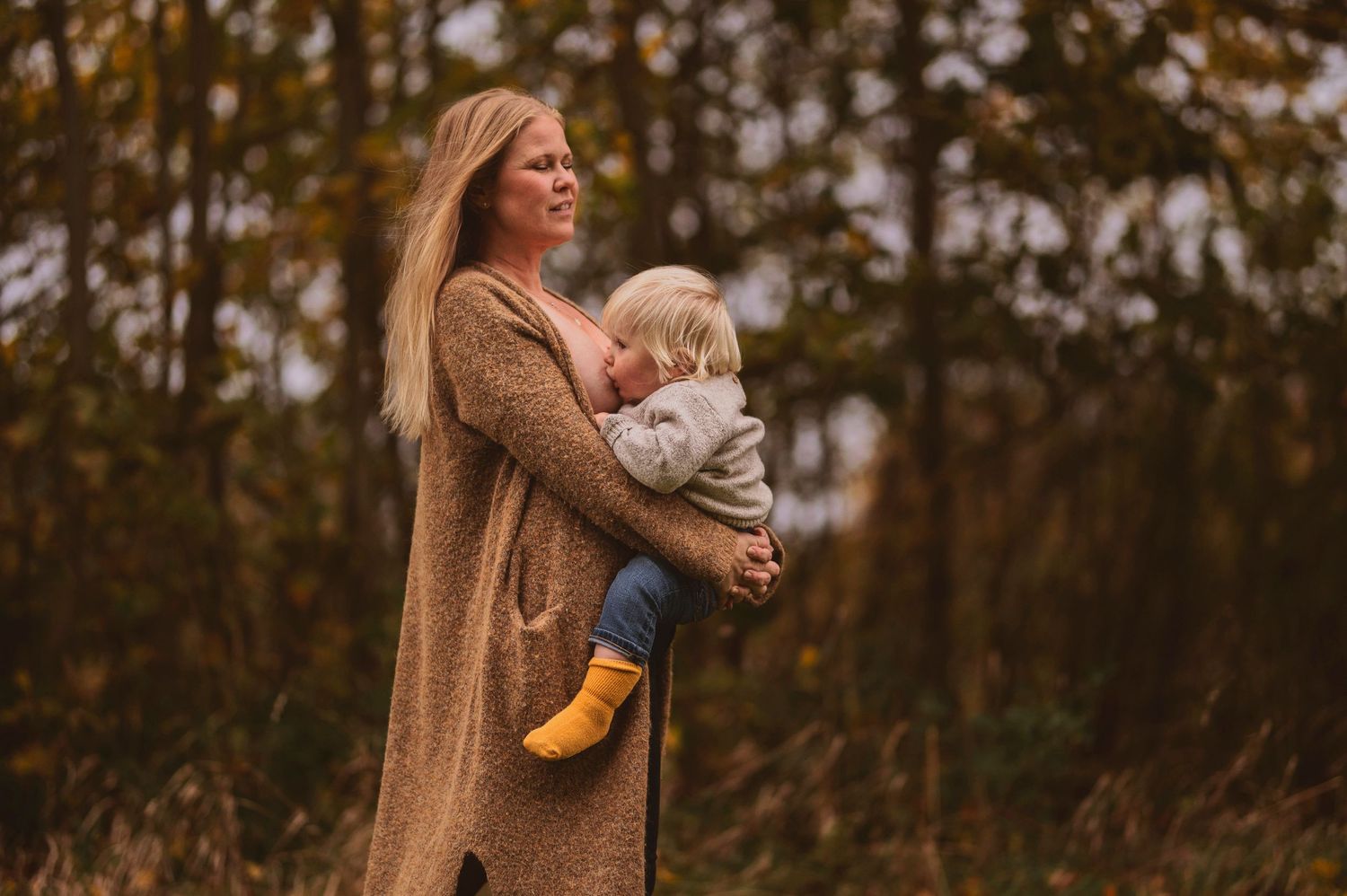 Adult wearing long brown coat embraces small child wearing yellow boots against autumn trees.