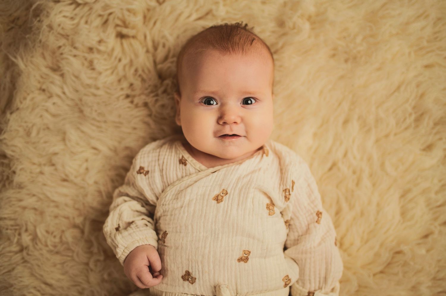Baby wearing cozy cream sweater with delicate pattern lying on soft fur backdrop.