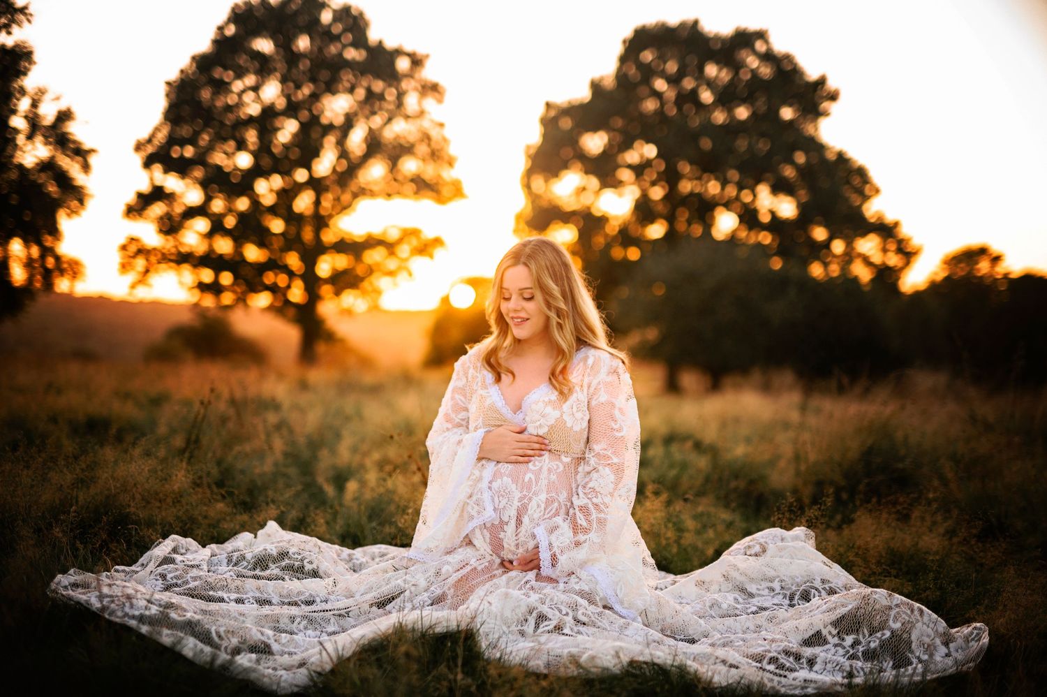 A lace maternity dress glows in golden sunset light as figure sits in grassy field with trees silhouetted behind.