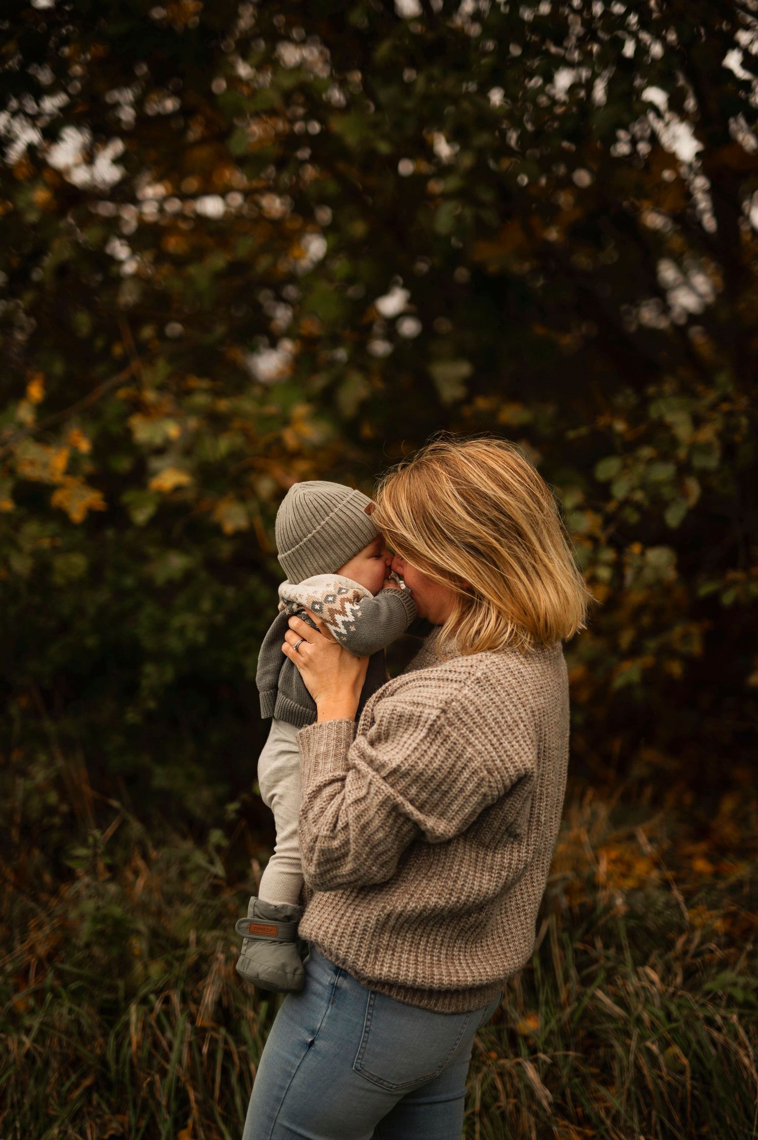 Tender moment captured outdoors with chunky knit sweaters and autumn foliage in background.