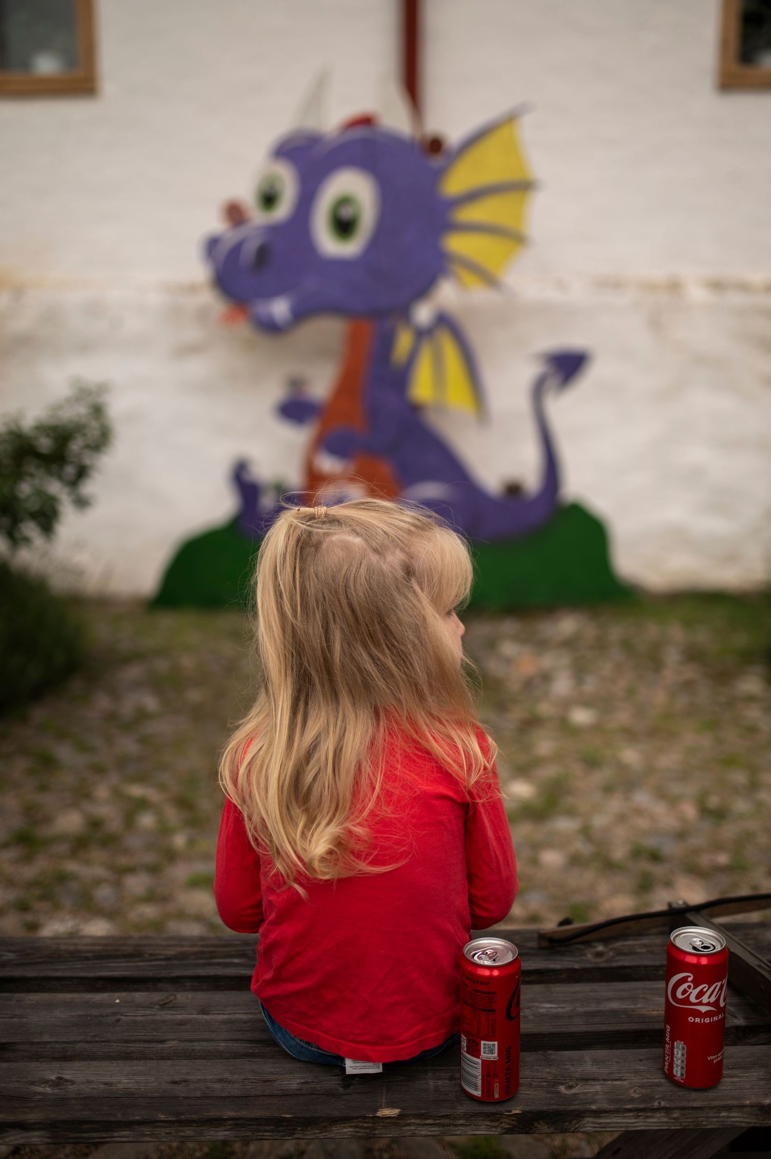 Child sits on wooden bench looking at colorful dragon mural with Coca Cola cans beside them.