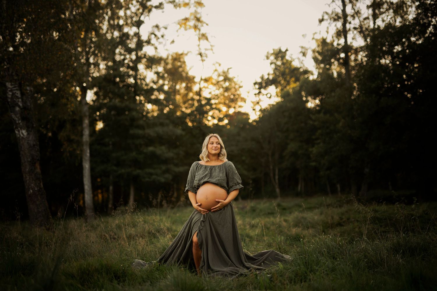 Ethereal maternity photograph captured in wooded meadow during golden sunset.