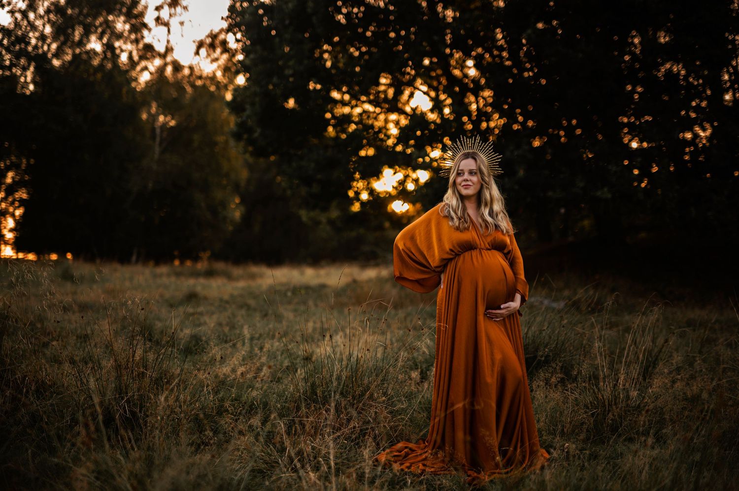 Silhouetted maternity portrait in flowing burnt orange dress against sunset-lit field.