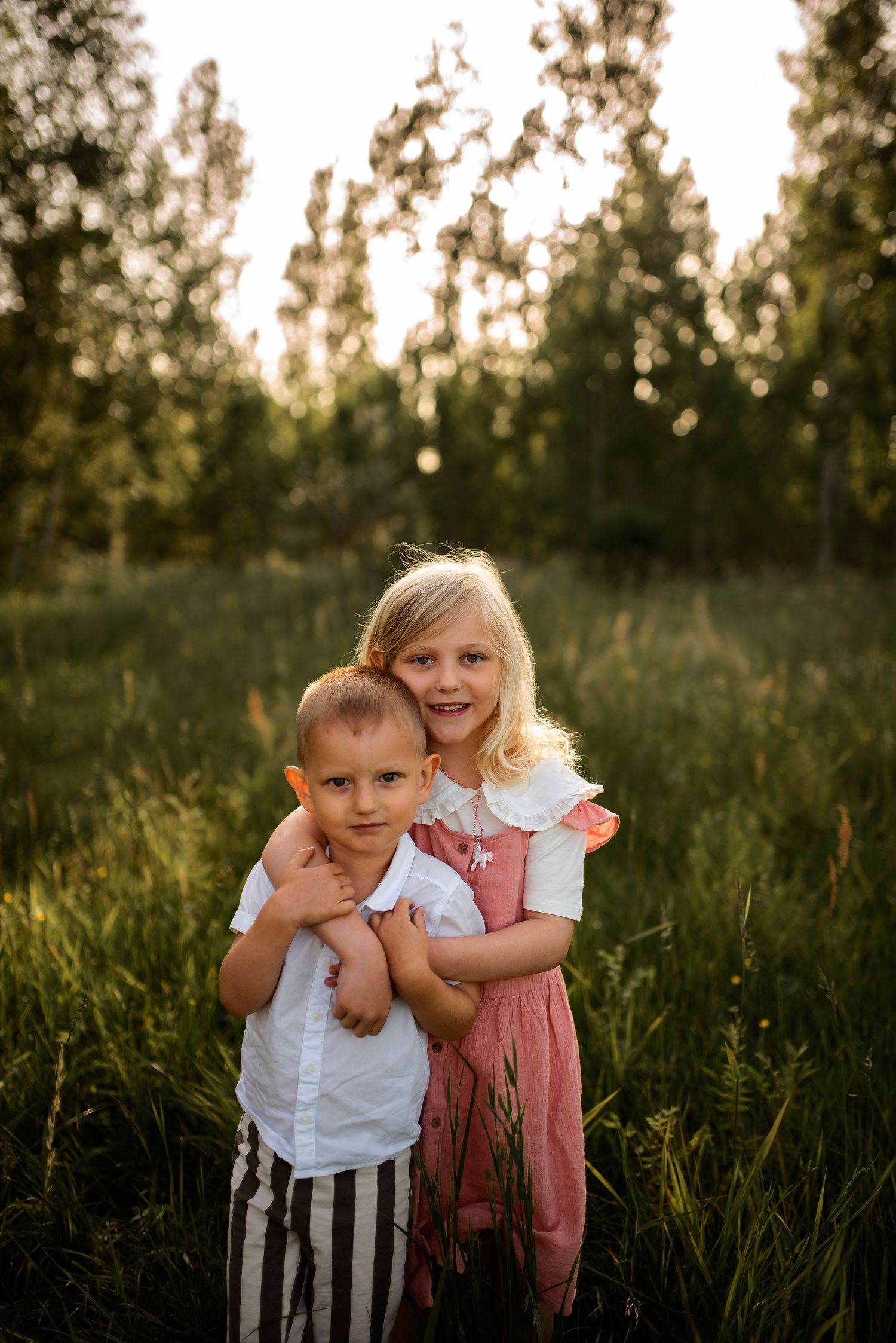 Two children hugging in tall grass during golden hour.