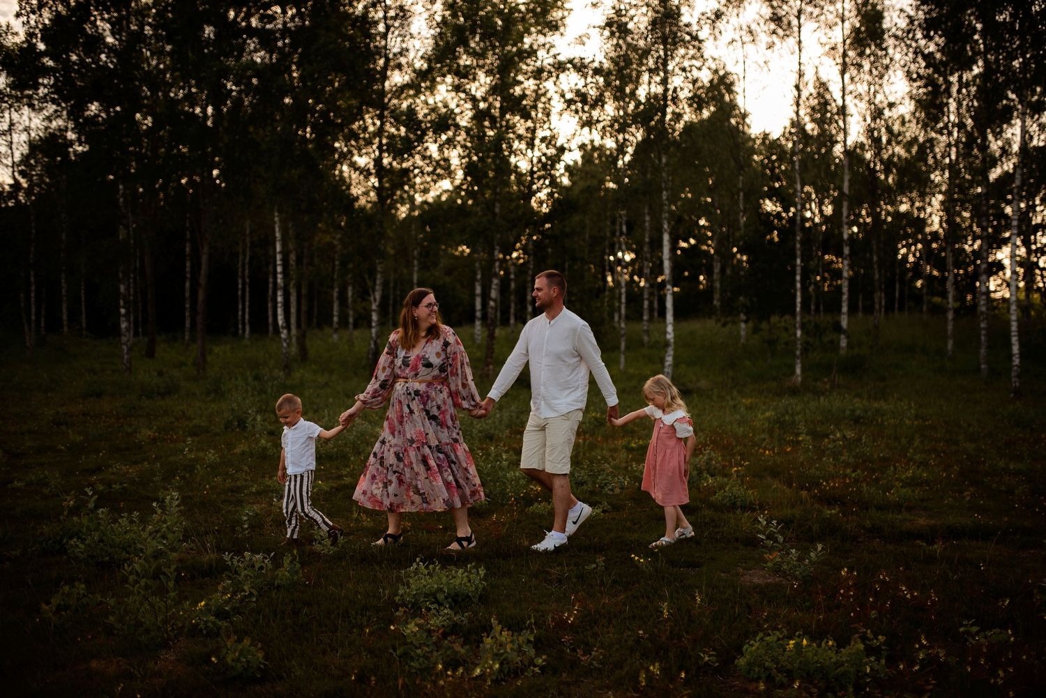Family walks hand in hand through field at dusk with forest backdrop.
