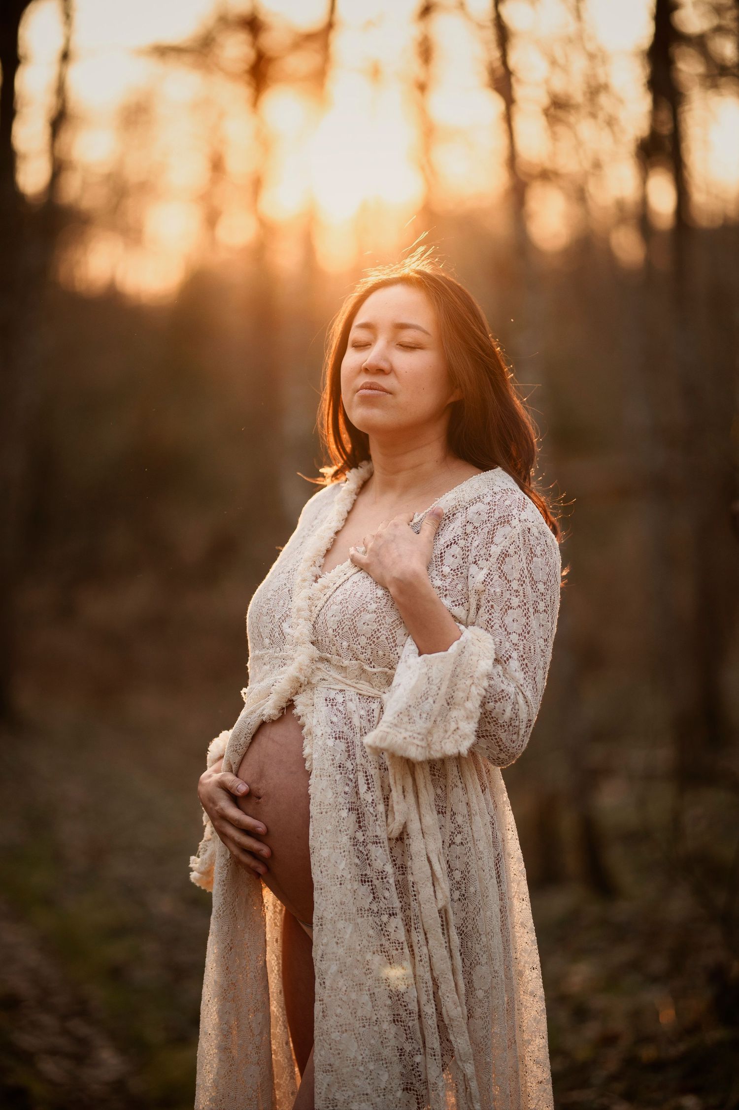 A serene maternity scene at sunset shows an expectant mother in a white lace dress touching her belly in a forest.