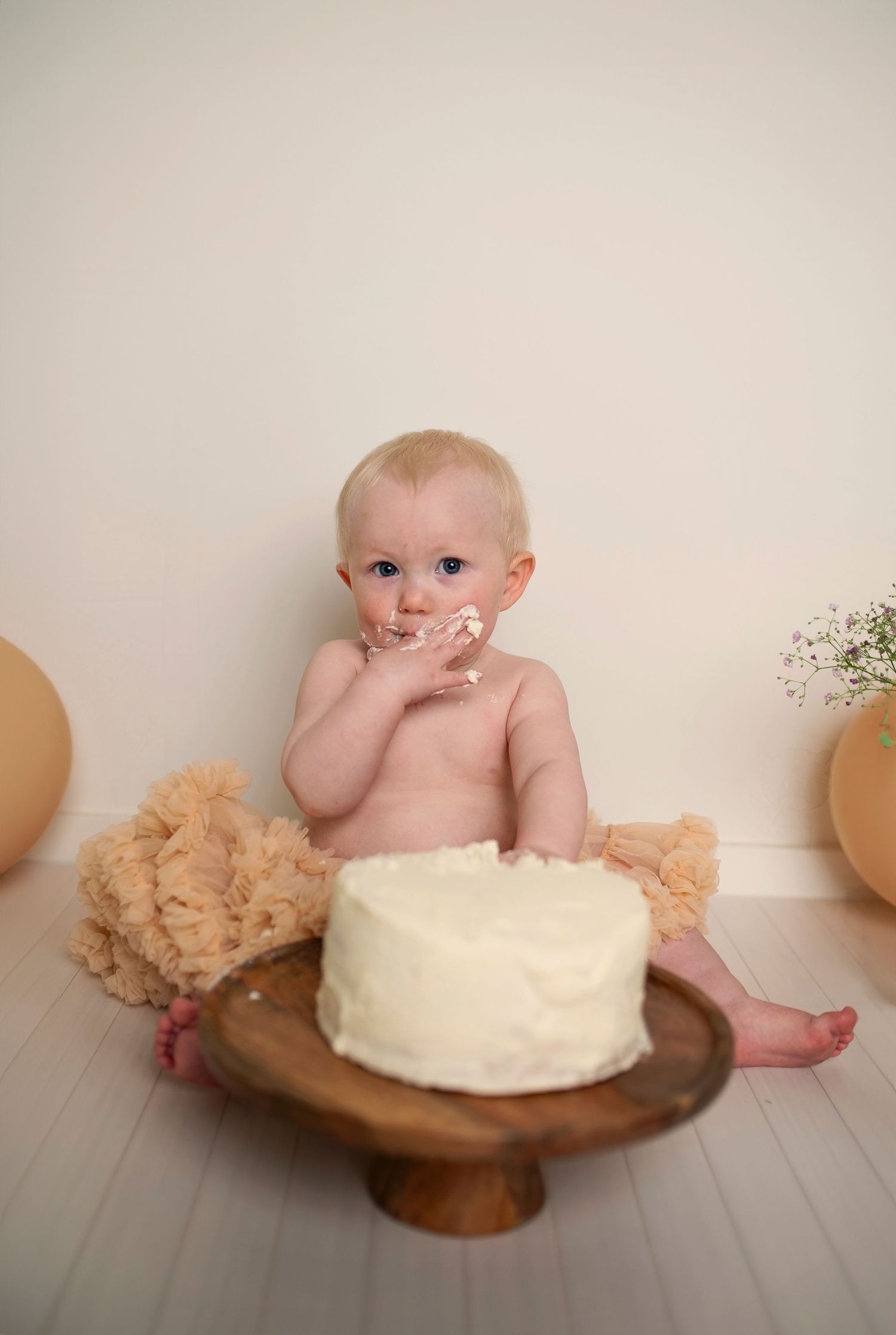 Baby reaching toward white cake on wooden pedestal with teddy bear and balloons in background.