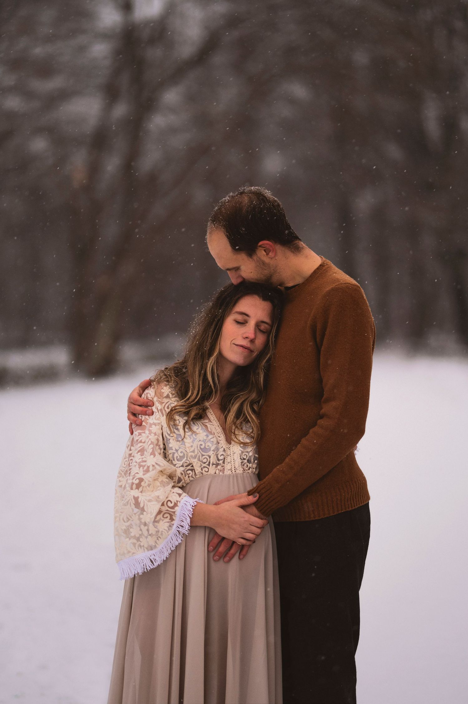 An intimate maternity portrait shows a couple embracing in the snow with the woman wearing a lace dress.
