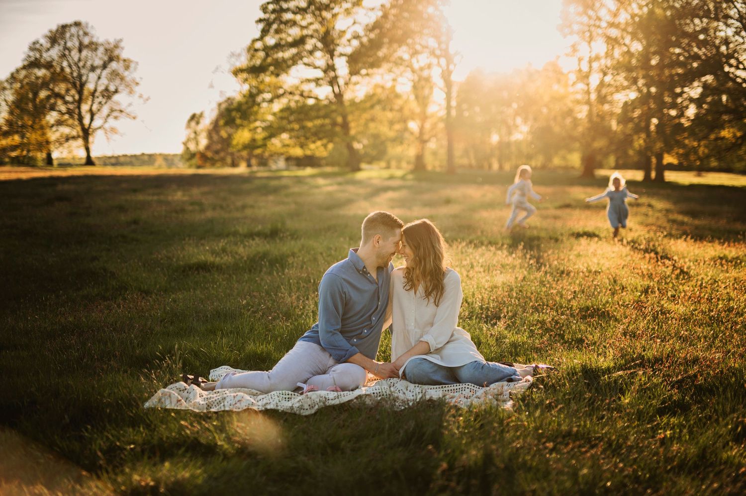 Couple sitting on blanket in park during golden hour with sun setting behind trees.