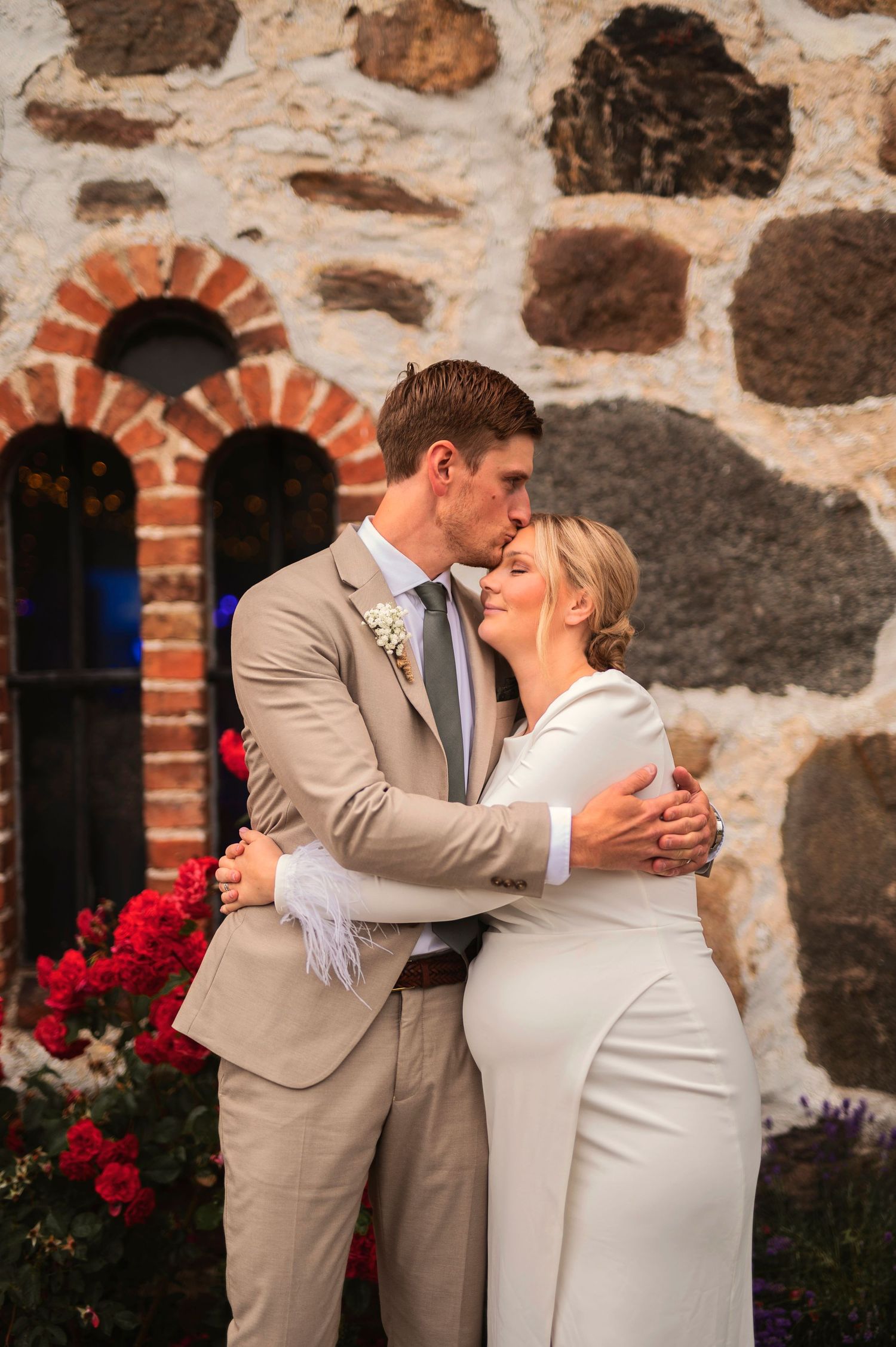 A romantic wedding couple embraces in front of a stone wall with red flowers and brick archways.