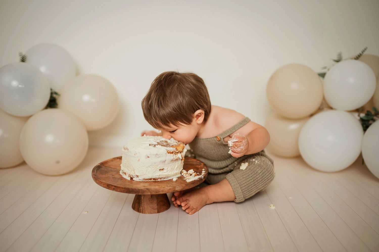 A baby in a white dress sits on the floor smashing and eating birthday cake surrounded by white balloons.