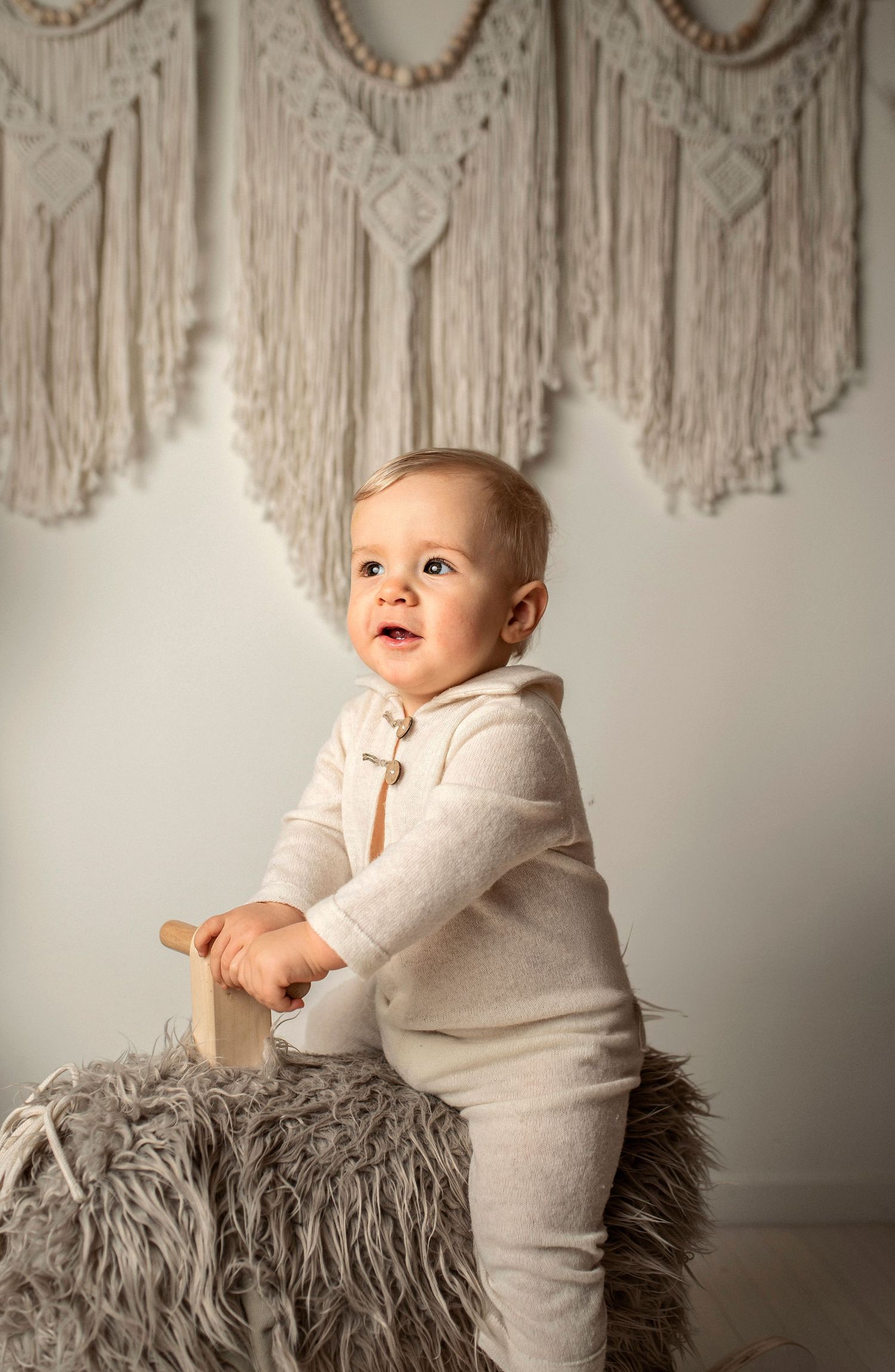 A small child in cream-colored outfit stands on a furry gray stool against a wall with macrame decorations.