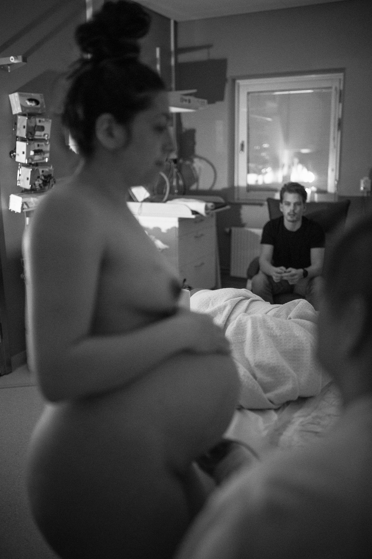 Black and white photo of nude pregnant woman in labor room with partner in background.