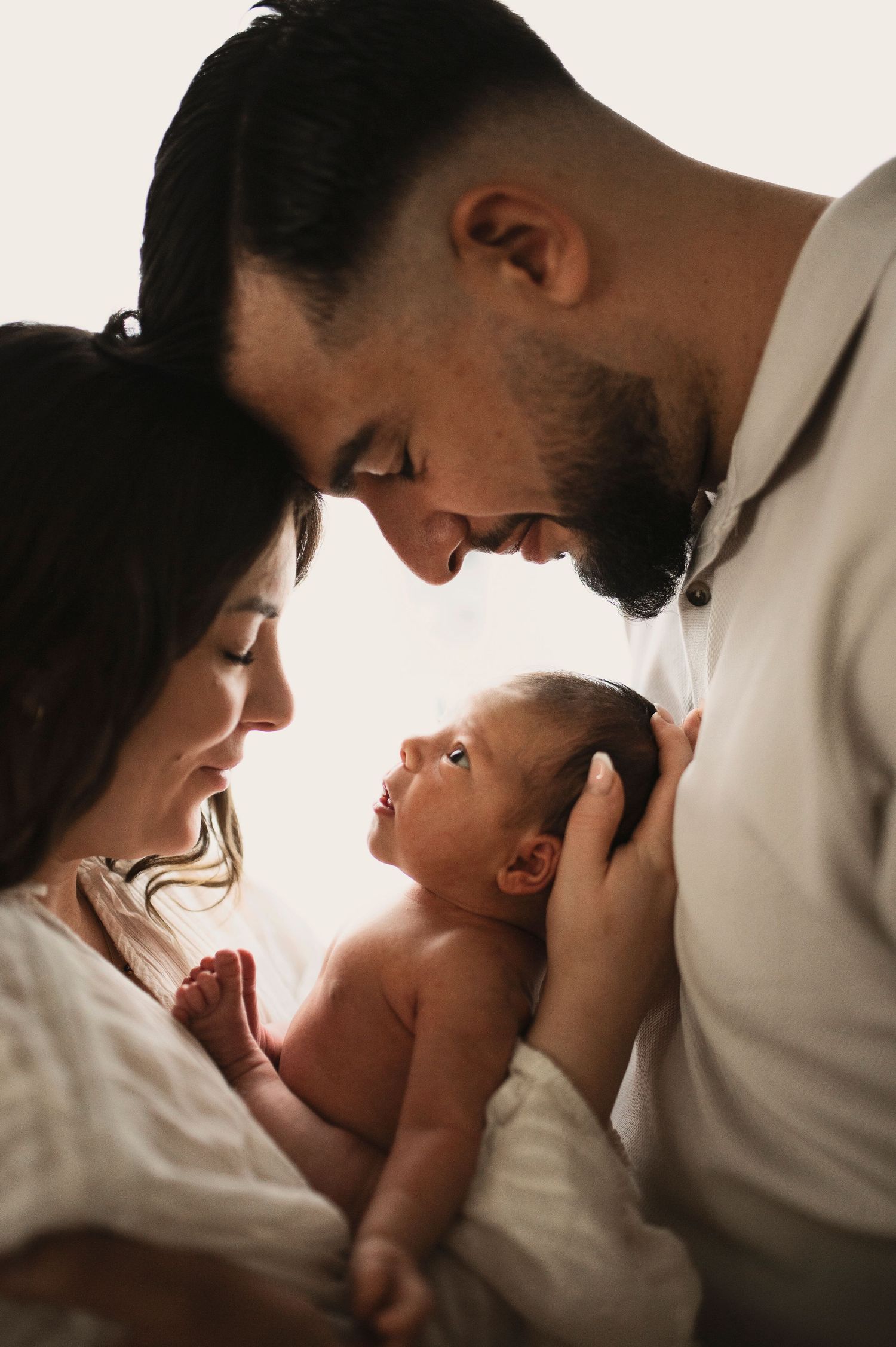 A tender family moment as parents lean in close to their newborn baby on white bedding in warm lighting.