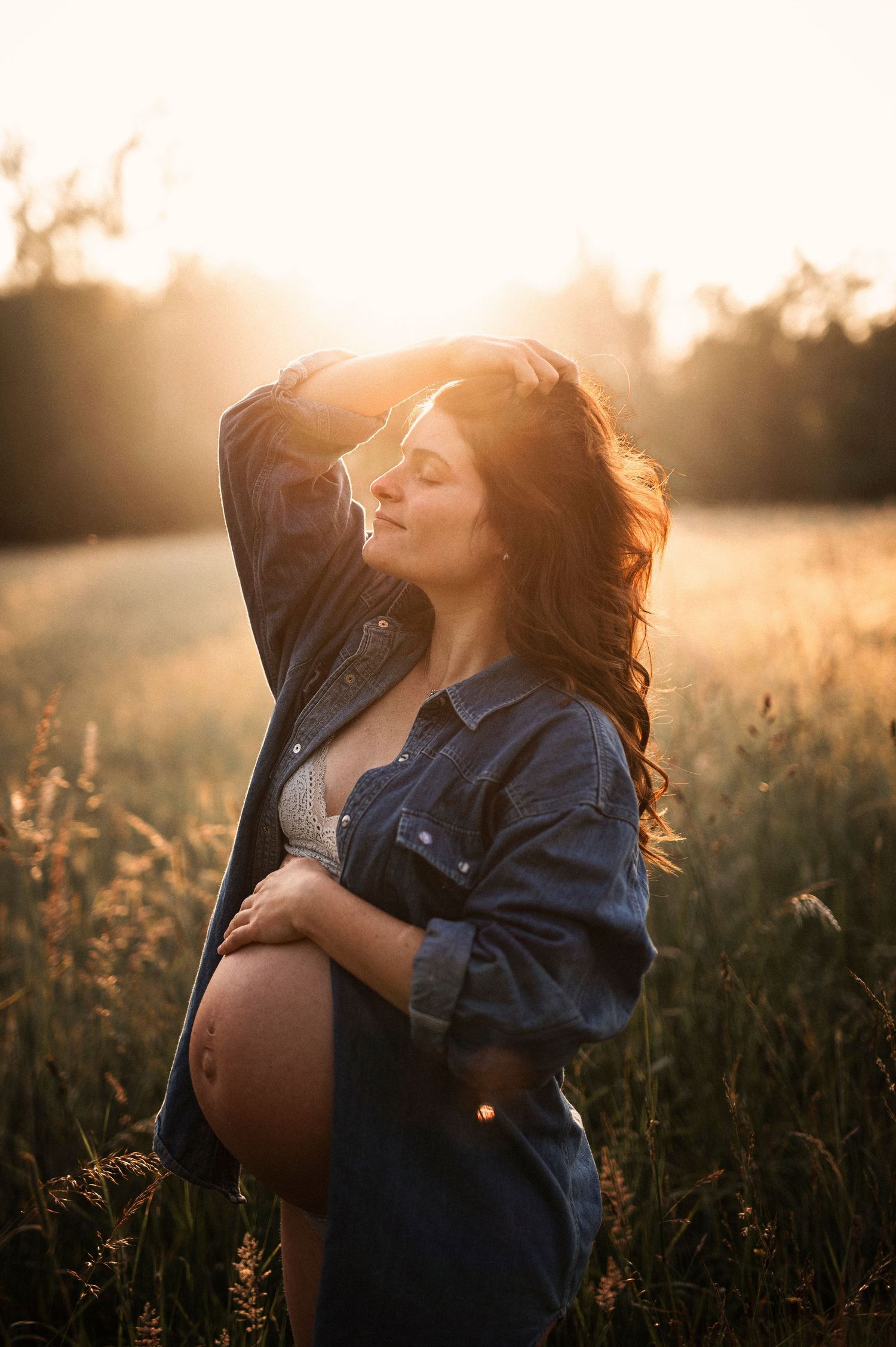 A maternity photo shows an expectant mother in a denim jacket standing in a golden field at sunset.