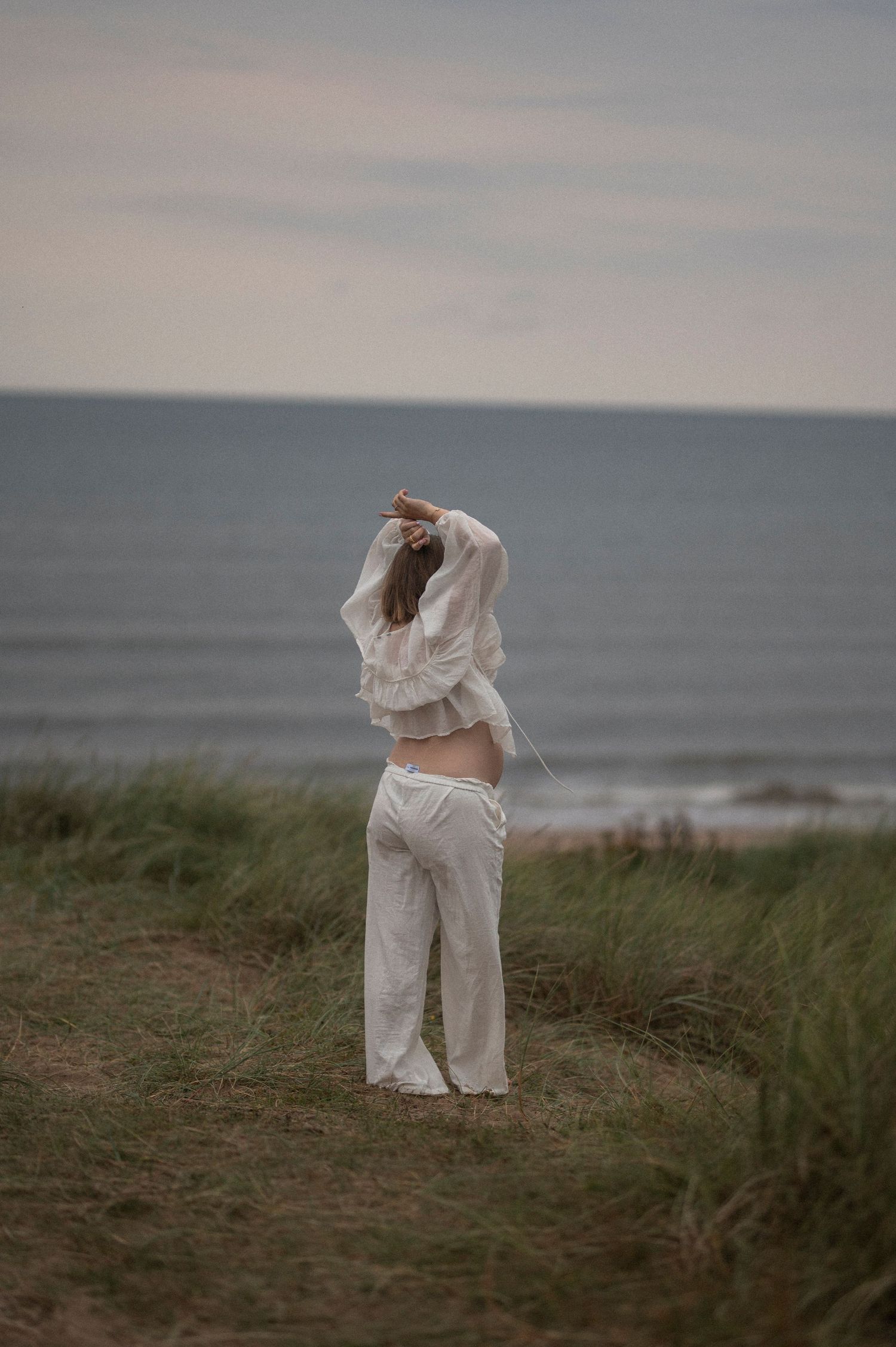 A silhouette stands contemplatively by the ocean in flowing white clothing.